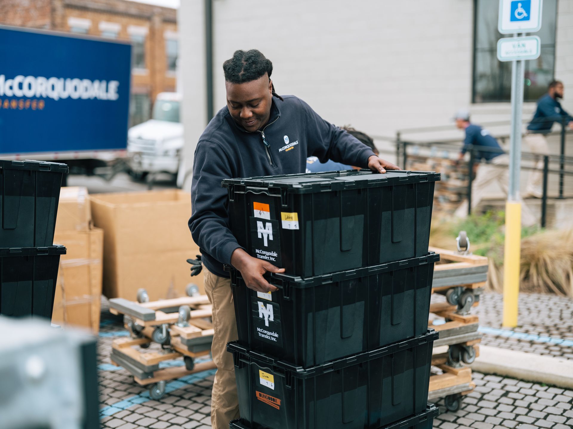 Man stacking black moving crates near a building with a McCORQUODALE truck in the background.