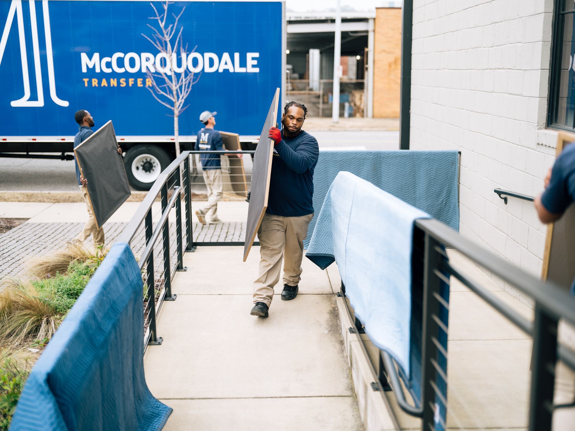 Movers unloading items from a moving truck onto a ramp, covered in blue padding.