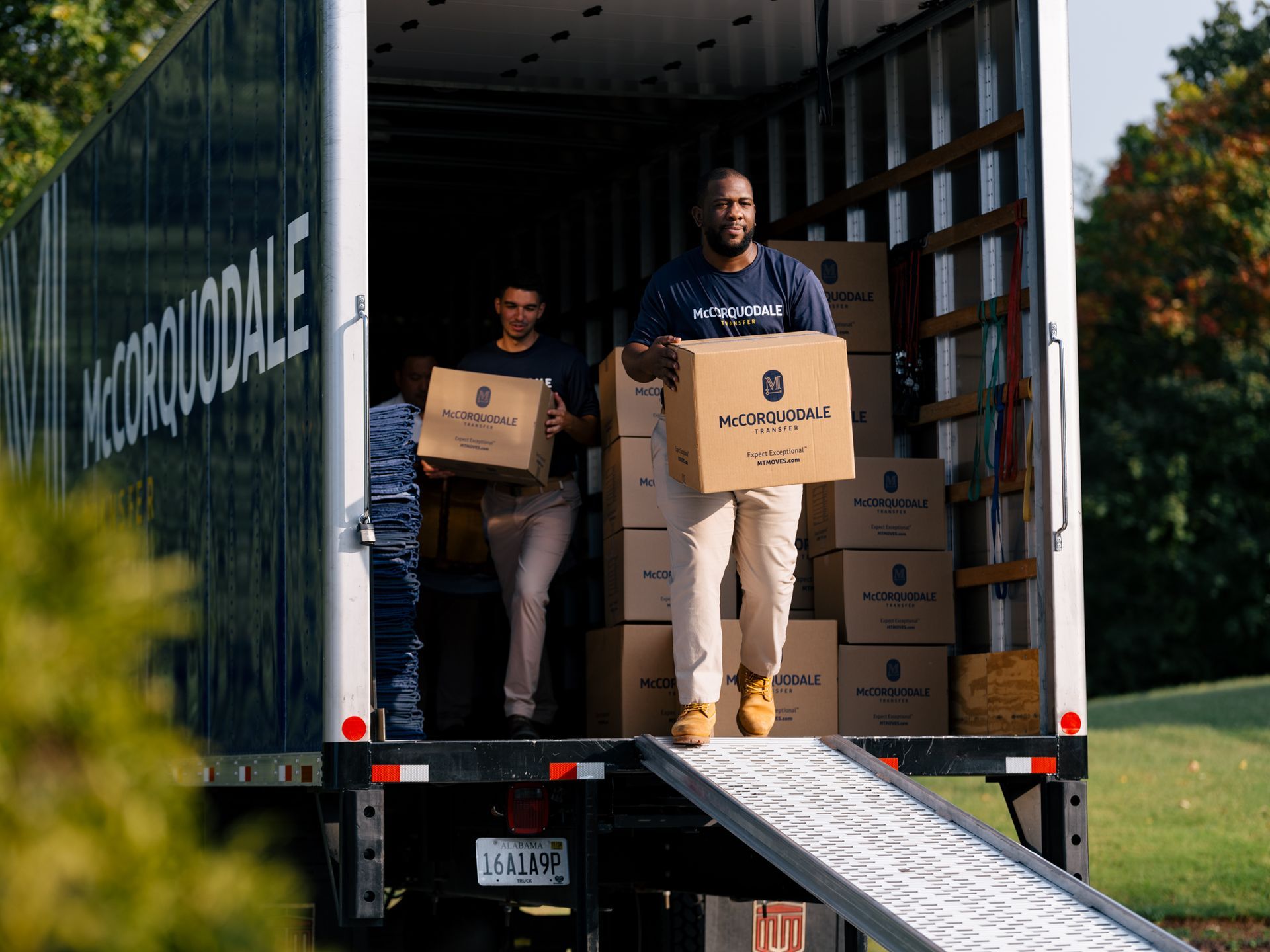 Men unloading boxes from a moving truck onto a ramp, outdoors.