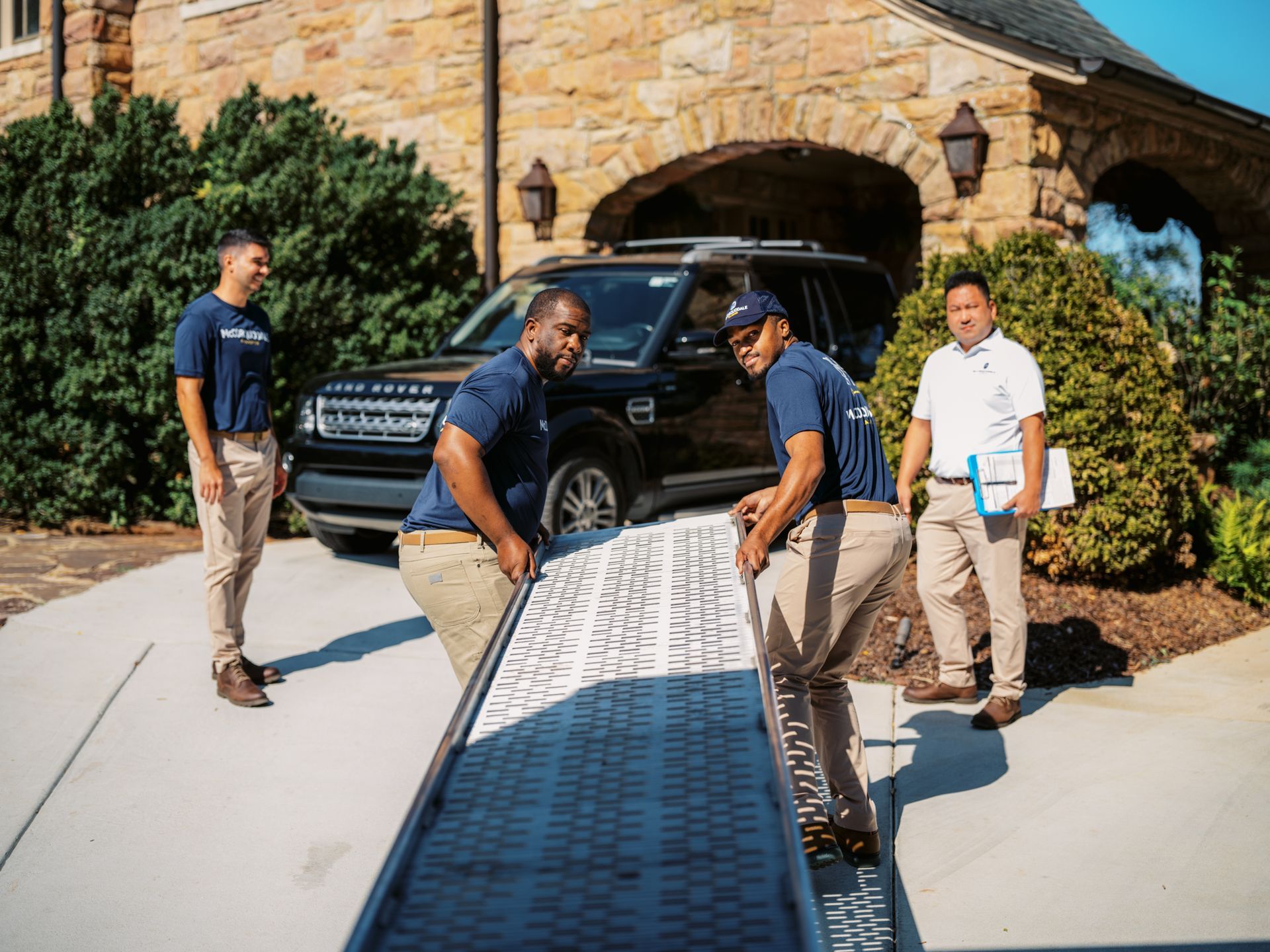 Movers loading a ramp into a black SUV parked in front of a stone house.