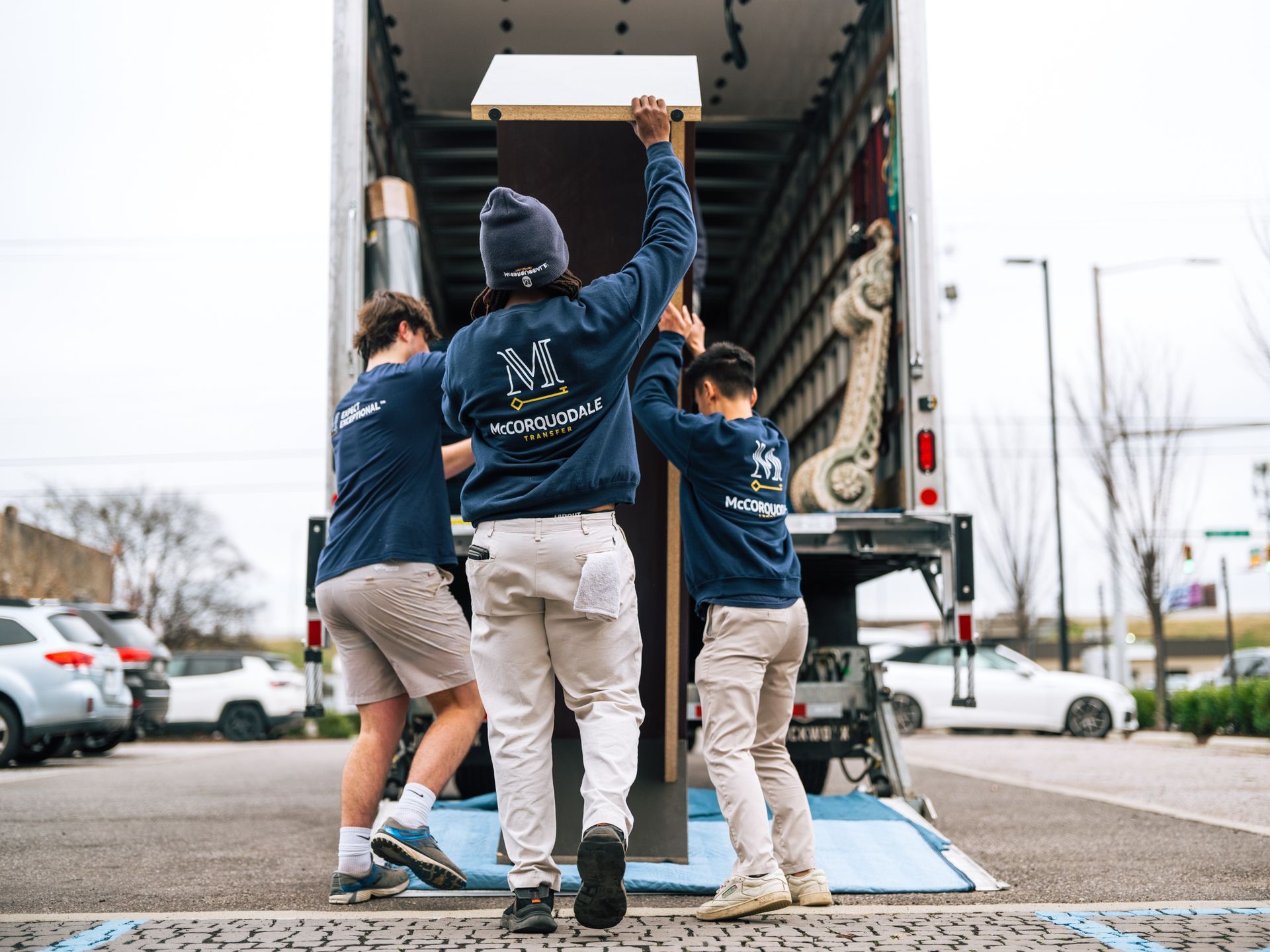 Three movers loading furniture into a truck. They wear matching blue shirts, outside in a parking lot.