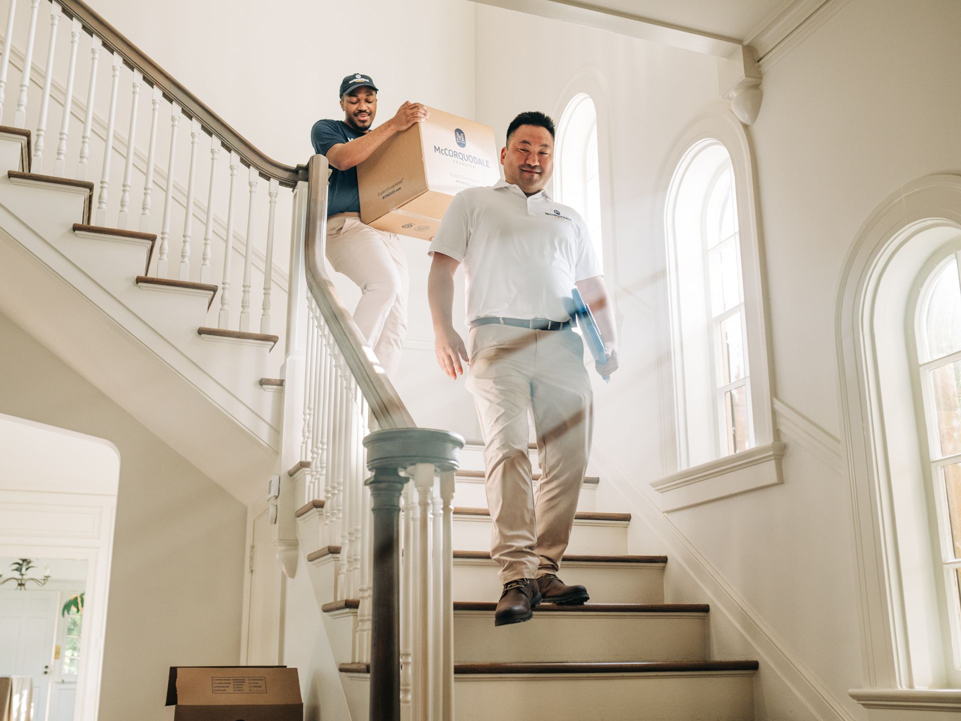 Two movers carrying a box down a bright staircase. One follows behind, the other ahead.