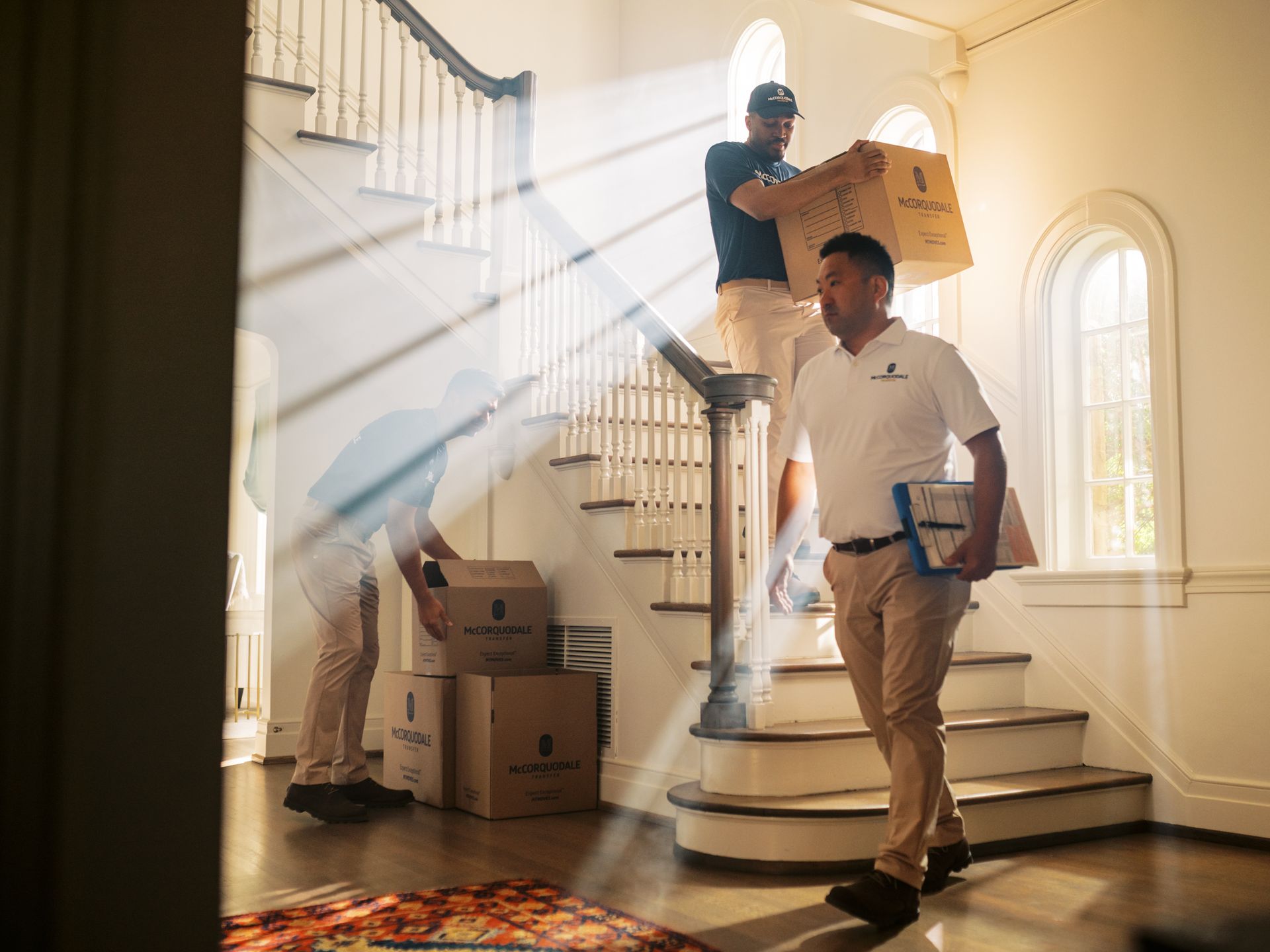 Movers carrying boxes up a staircase in a well-lit house. One man leads with a clipboard. Sunlight streams through a window.