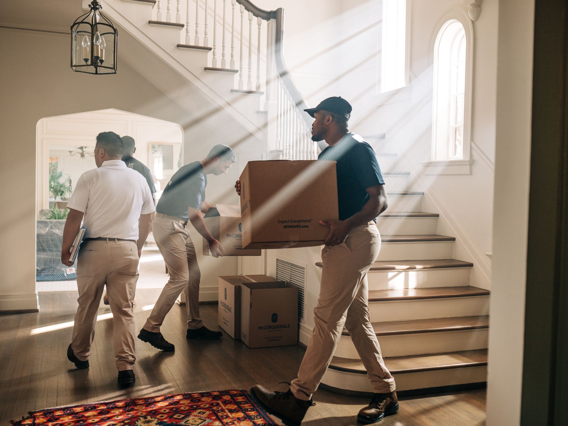 Movers carrying boxes through a brightly lit foyer with a staircase.