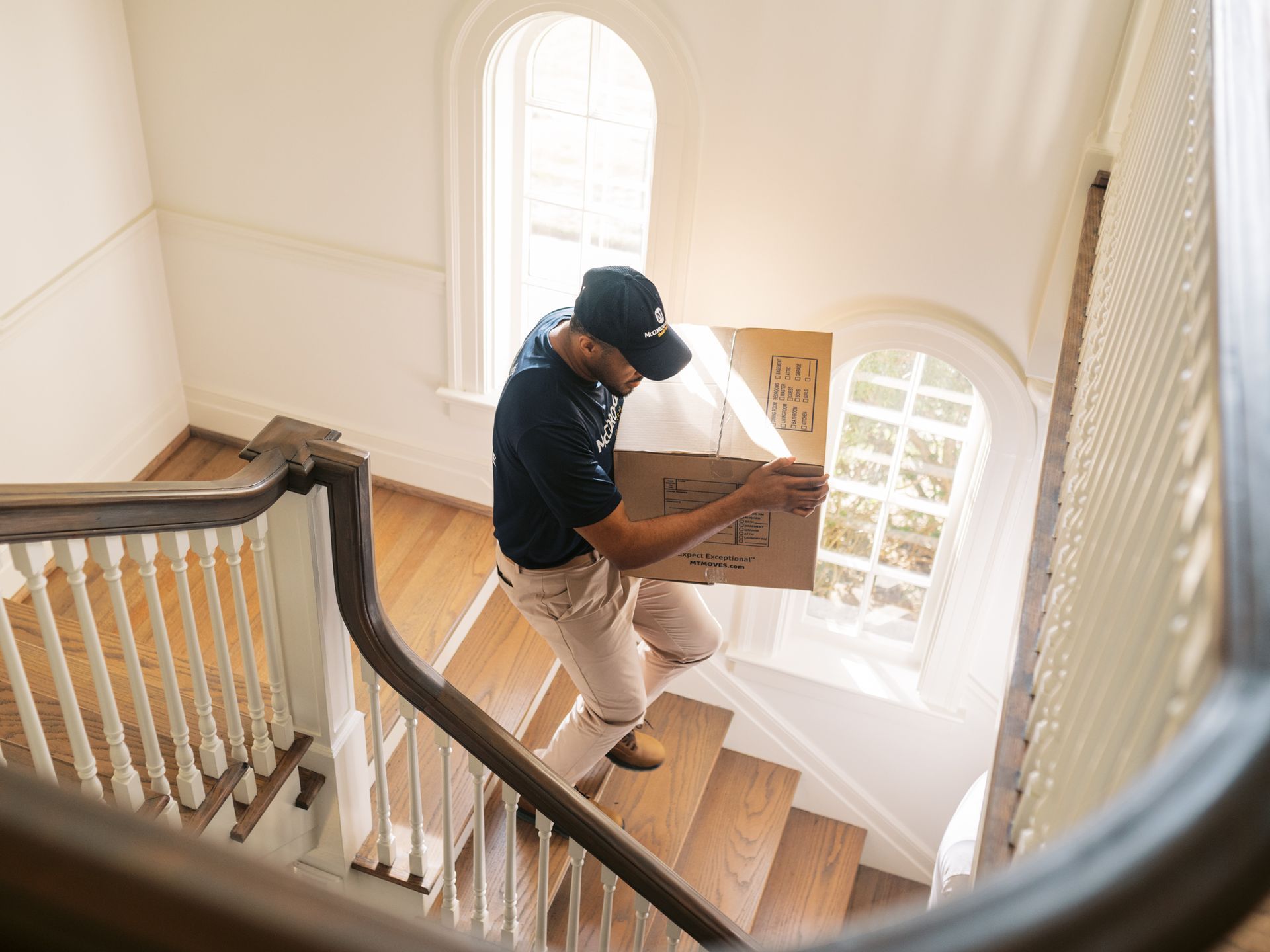 Man carrying a cardboard box down wooden stairs in a house with arched windows.
