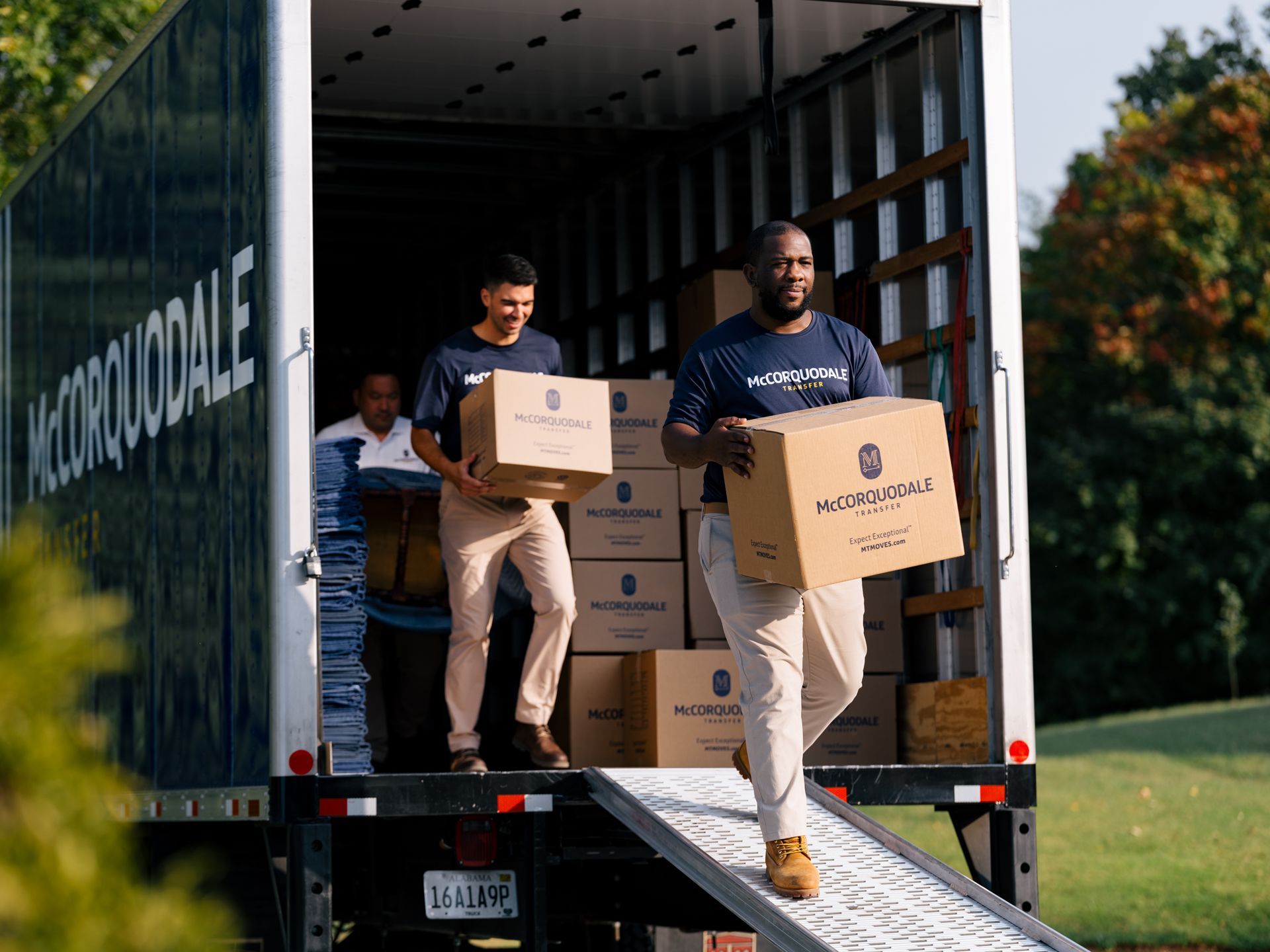 Movers unload boxes from a truck on a ramp. Outdoors with green grass and trees.