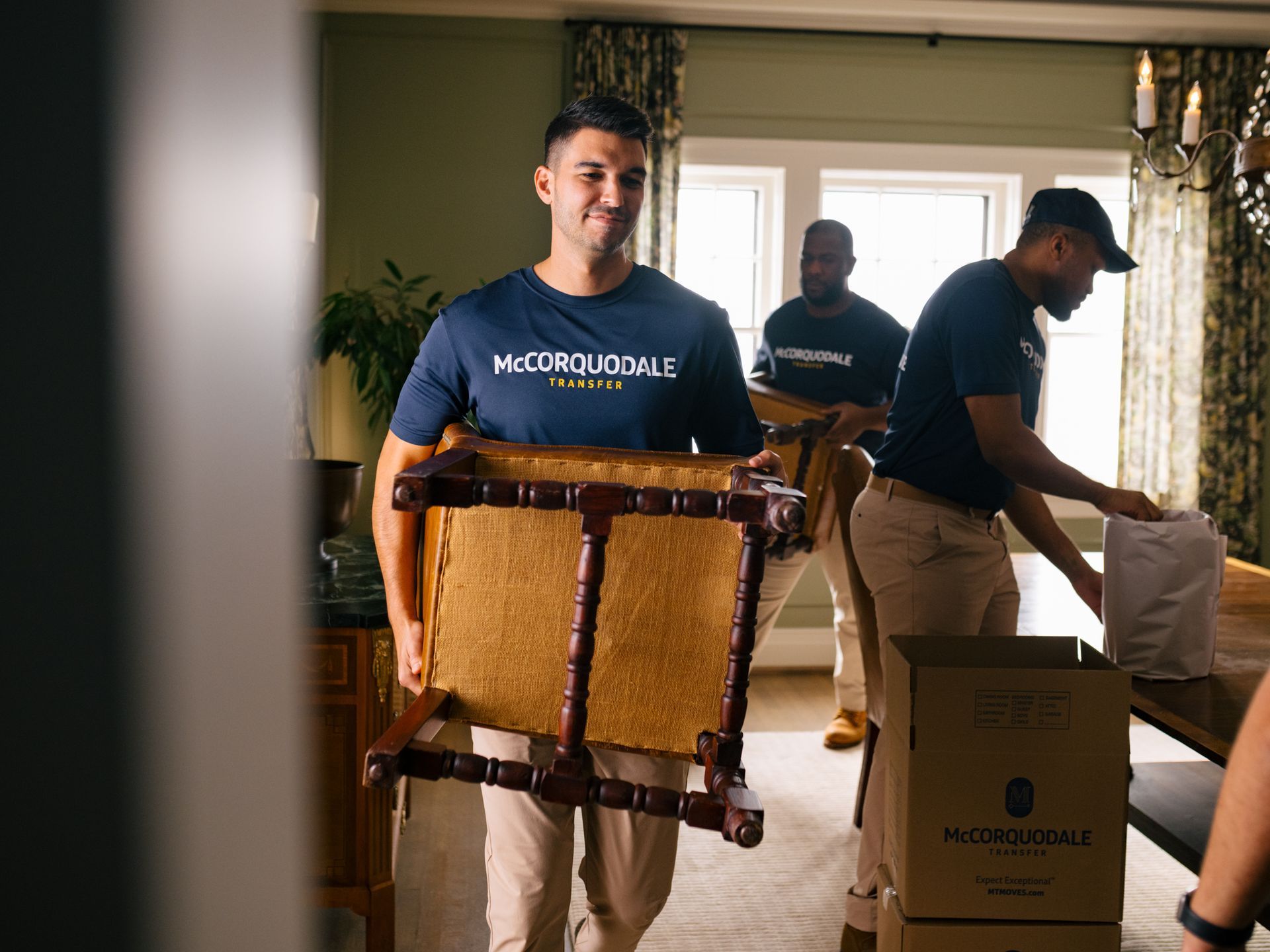 Movers carrying furniture inside a home. Man smiles while holding a chair; others pack boxes.