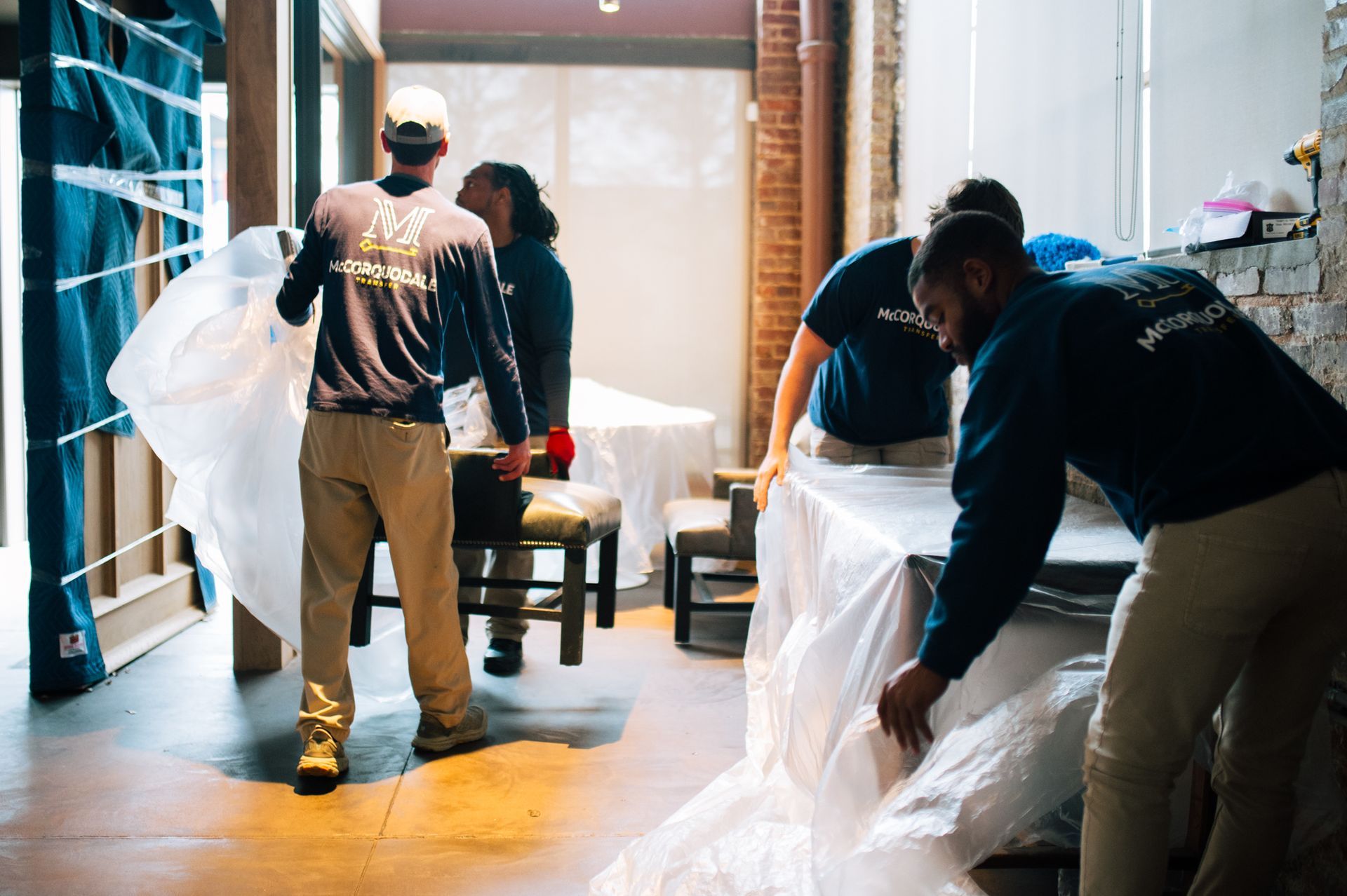 Movers wrapping furniture in clear plastic in a room with exposed brick and wooden accents.