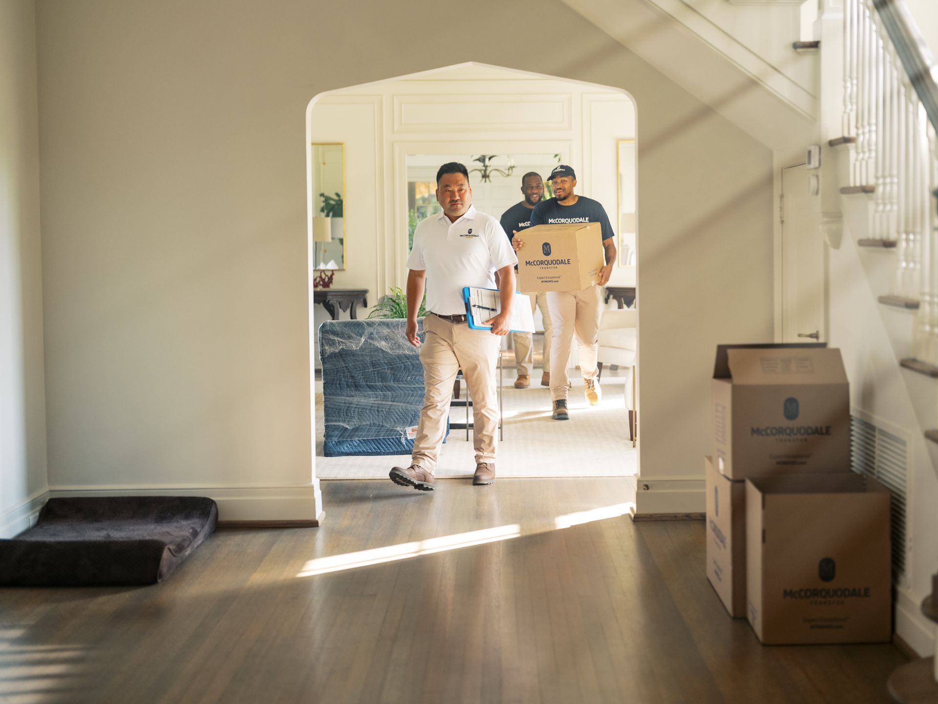 Movers carrying boxes through a doorway; one man leads the way. Sunlight streams in.