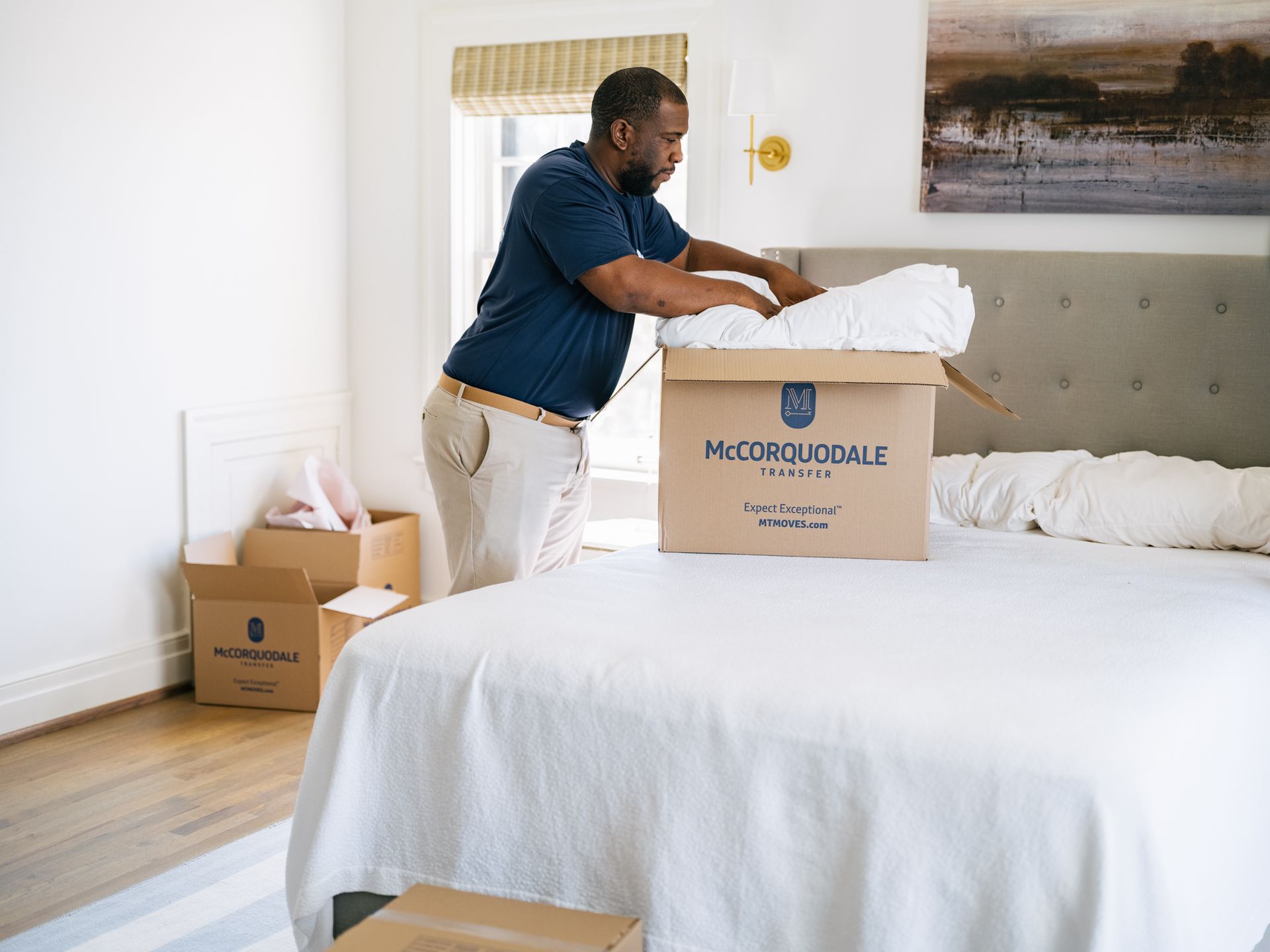 Man unpacking bedding from a moving box on a bed in a bedroom.