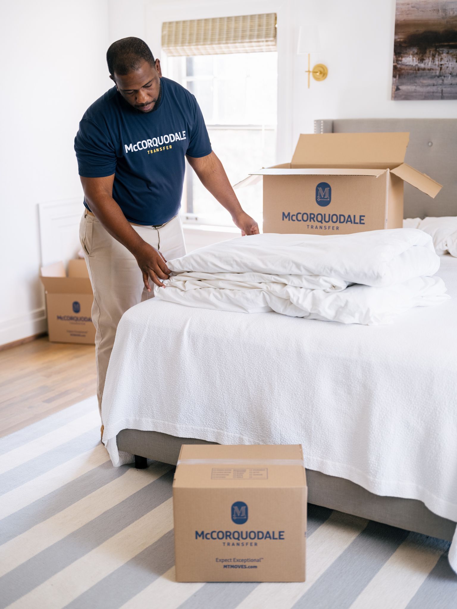 Man packing bedding from a bed into a labeled moving box in a bedroom.