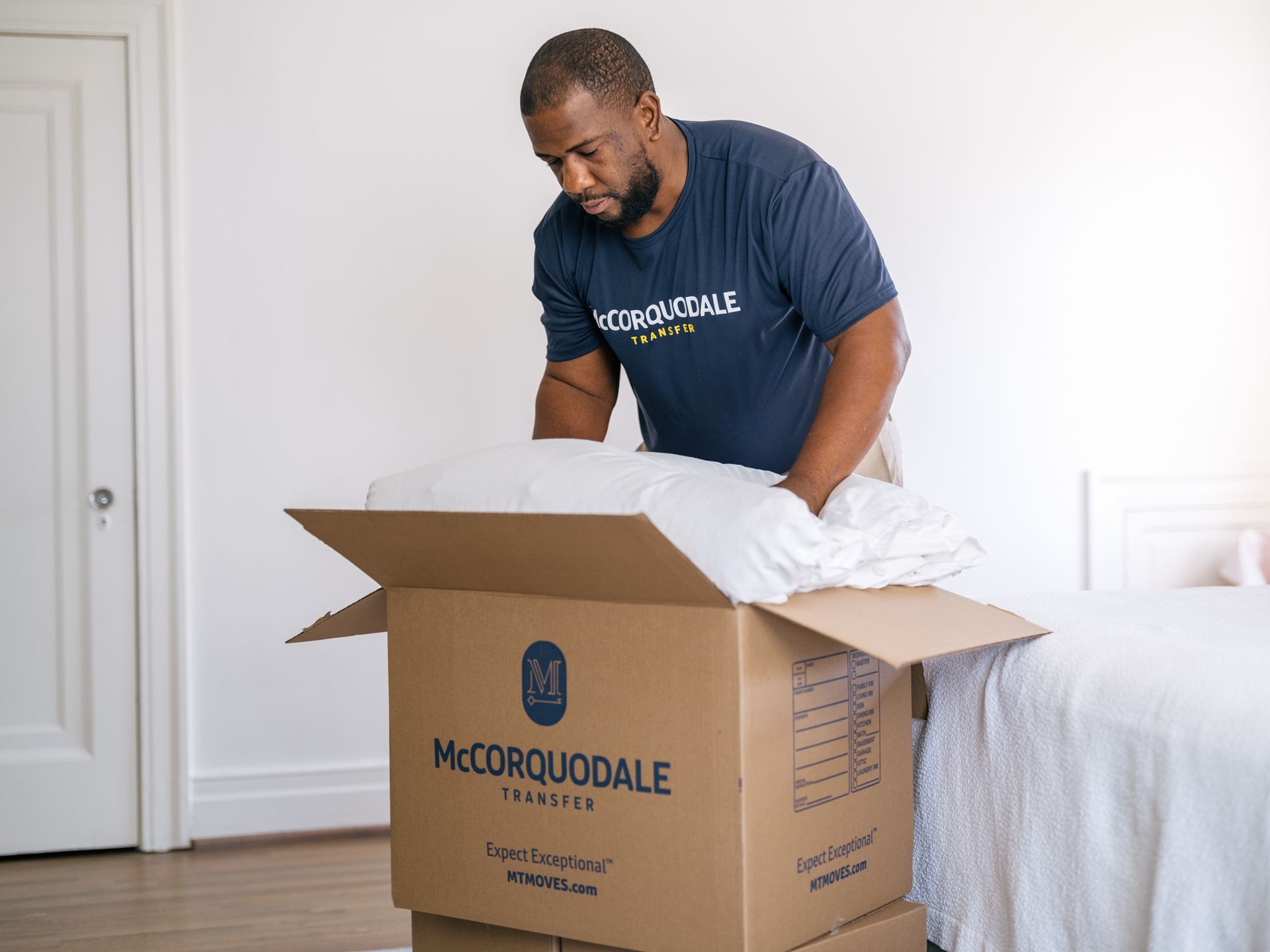 Man packing items into a cardboard box indoors; a bed and door are visible.