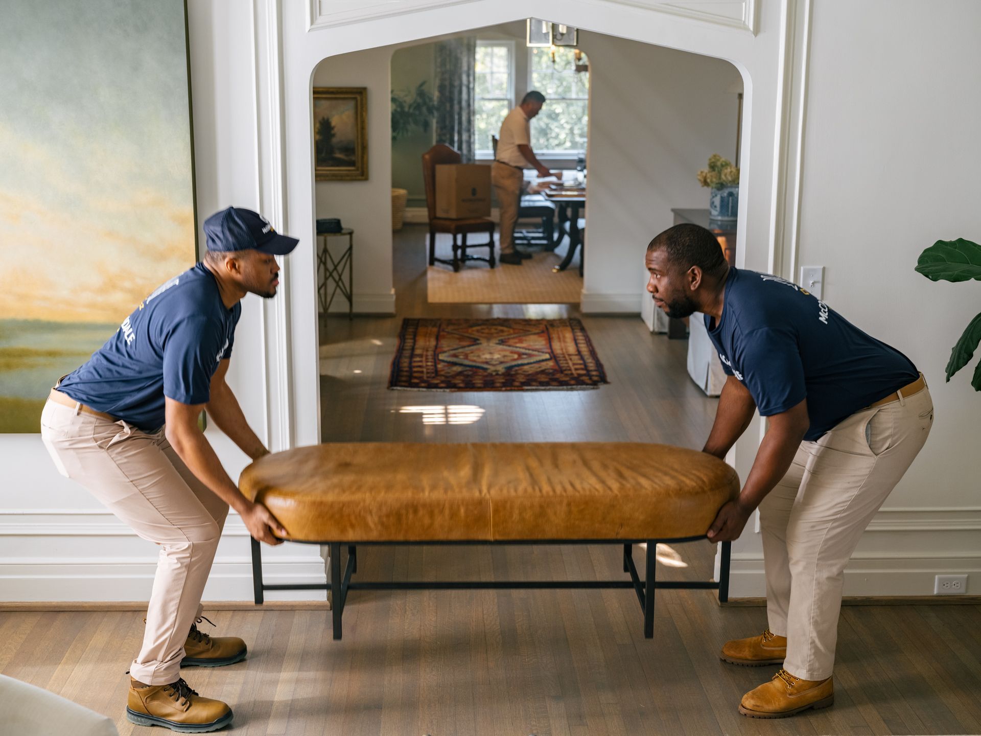 Two movers carrying a brown leather bench through a home's doorway. One man is visible in the distance.