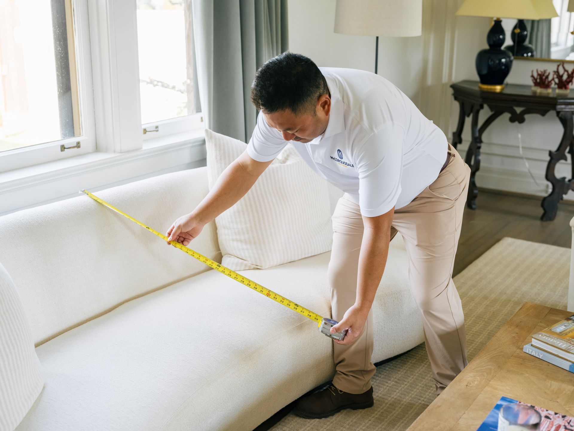 Man in white shirt measures a white sofa in a living room with a measuring tape.