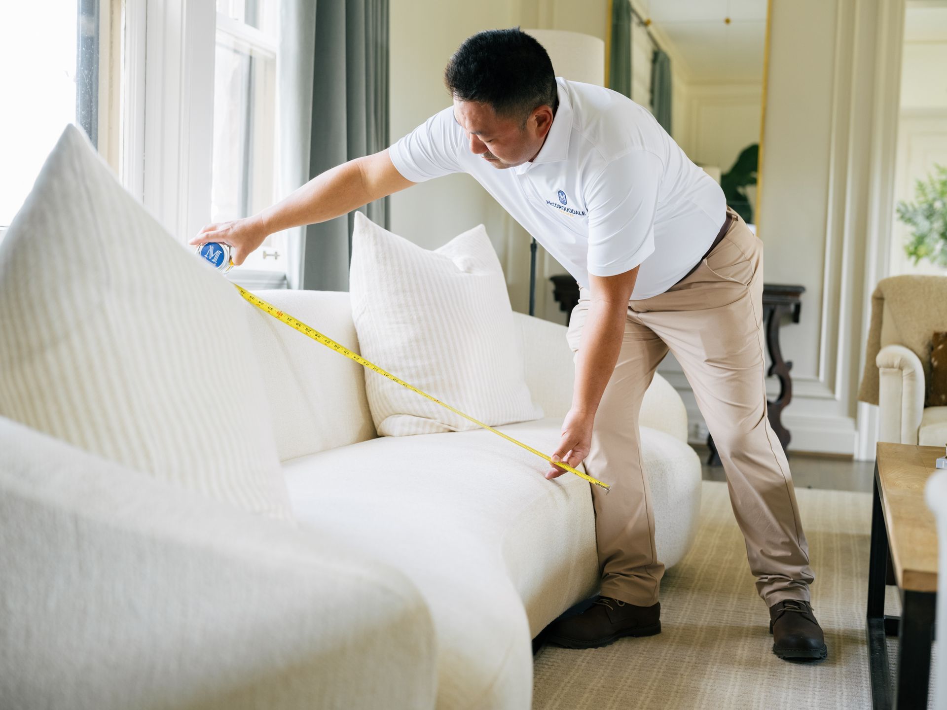 Man in white shirt measures a white sofa in a living room with a measuring tape.