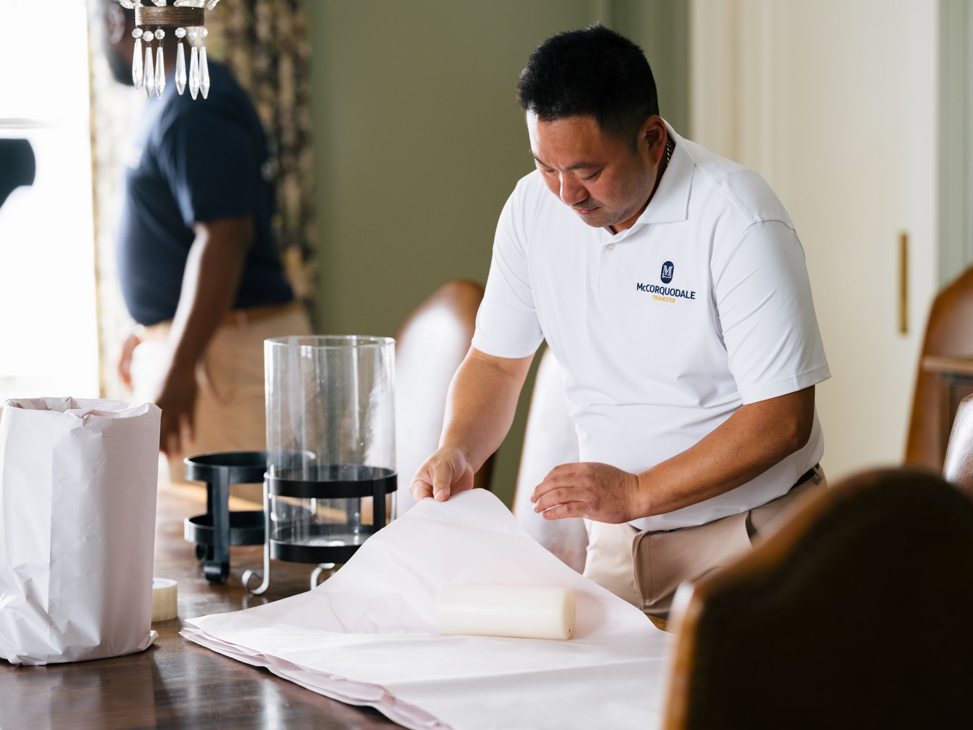 Man in white shirt wraps item on a dining table; another person in background.
