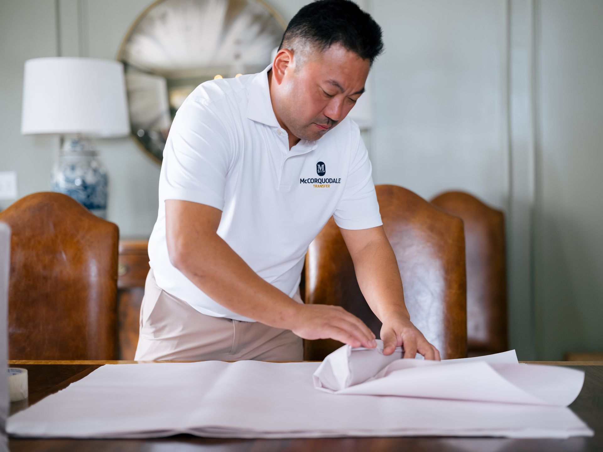Man in white shirt wraps item in paper on a table. Interior setting, neutral colors.