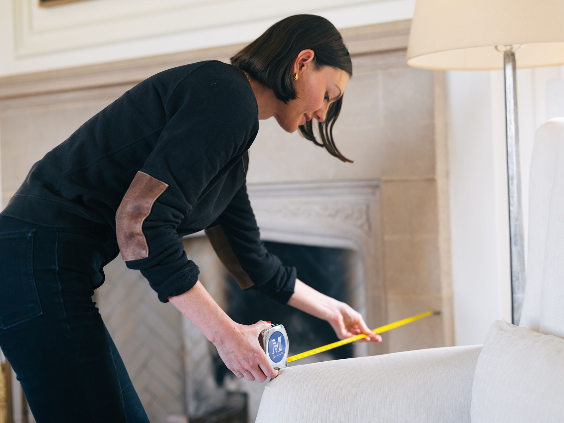 Woman in black measuring white couch in a living room near a fireplace and lamp.