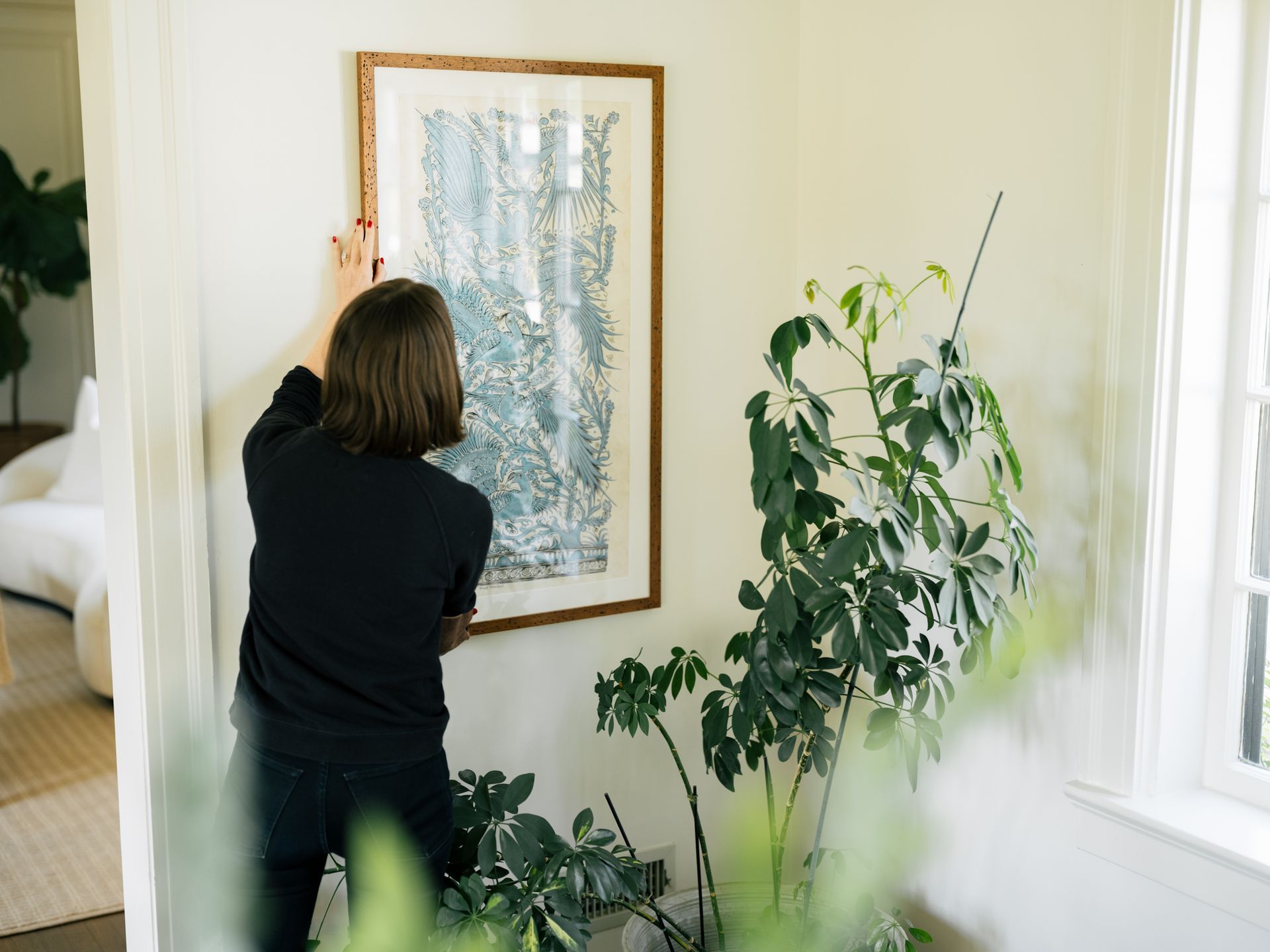 Person hangs art on a light-colored wall, surrounded by plants near a window.