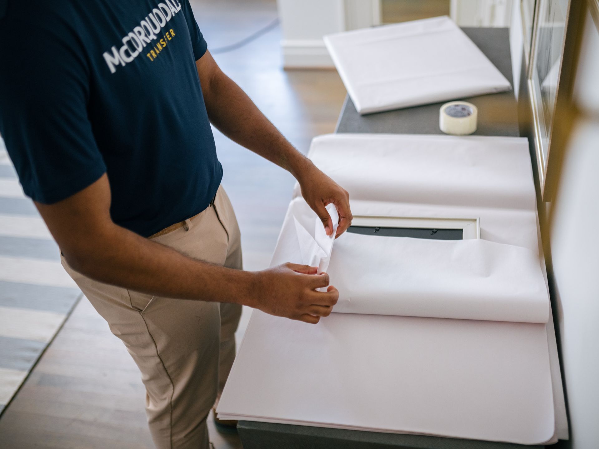Person wraps a rectangular item in white paper on a table, preparing for transport.
