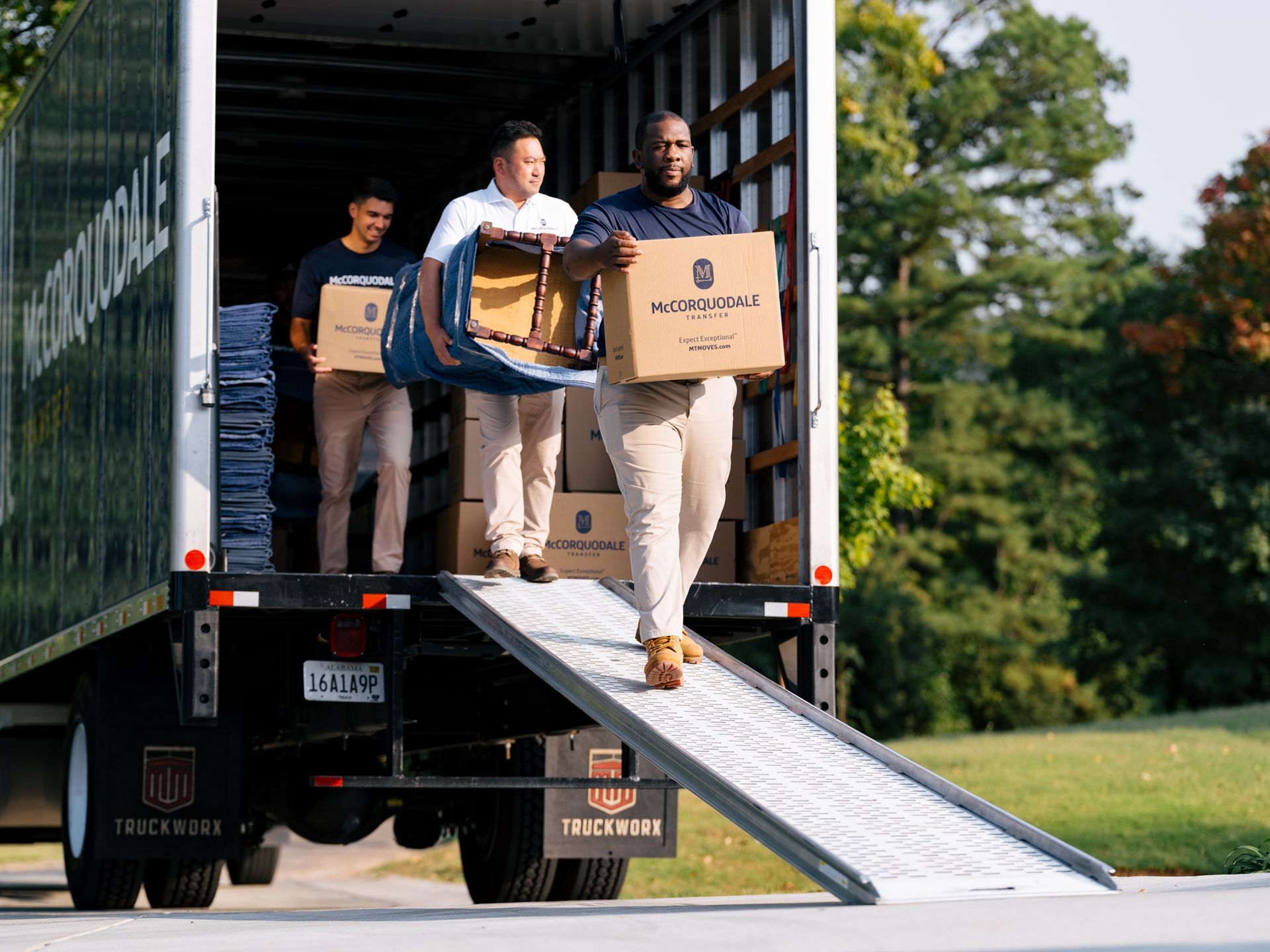 Three people loading a moving truck with boxes and furniture. Ramp extends from the truck to the ground.