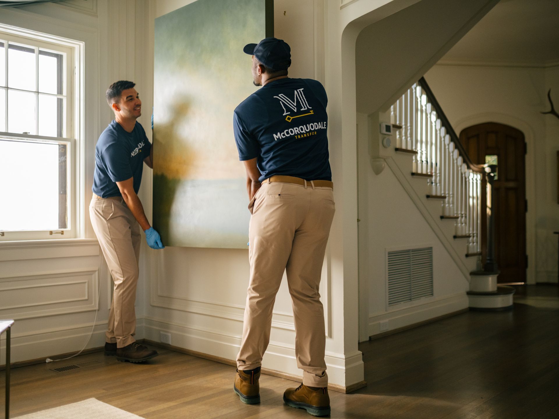 Two people wearing uniforms carry a large painting in a room with a staircase.
