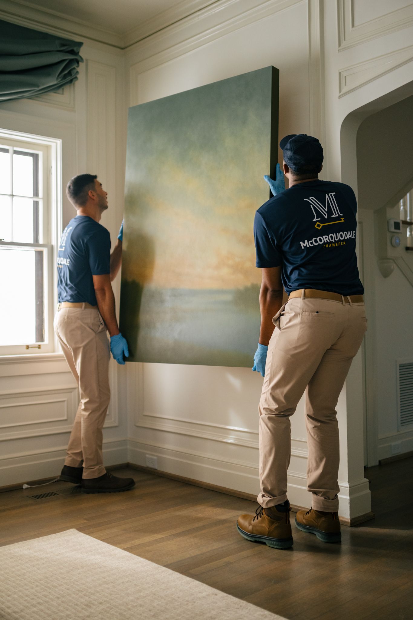 Two people in matching uniforms wearing gloves lift a large painting on a wall.