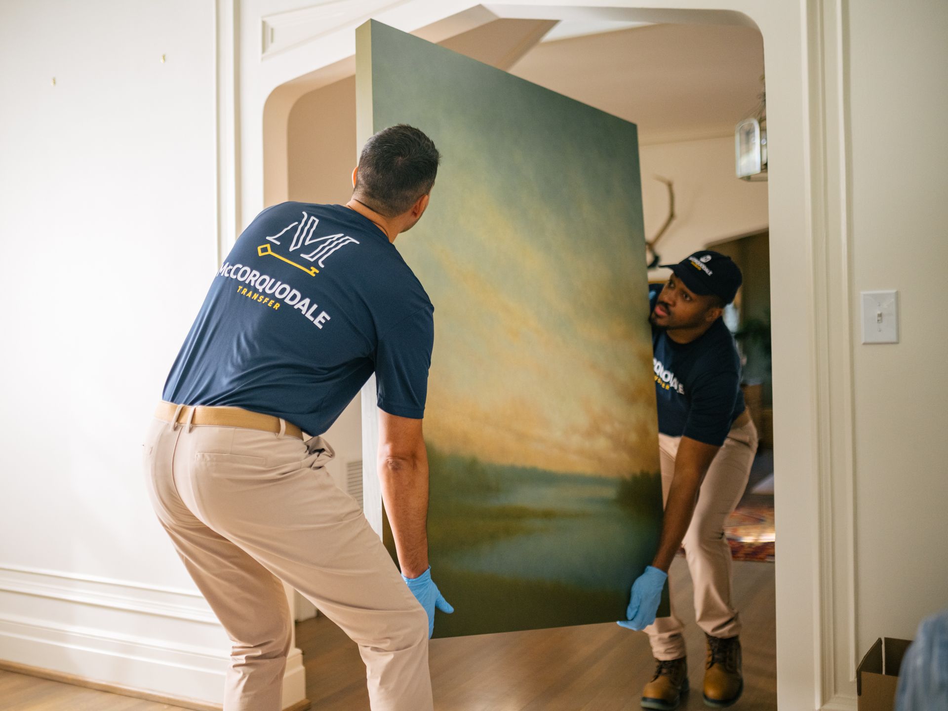 Two people carrying a large painting through a doorway. They wear blue shirts, tan pants, and gloves.