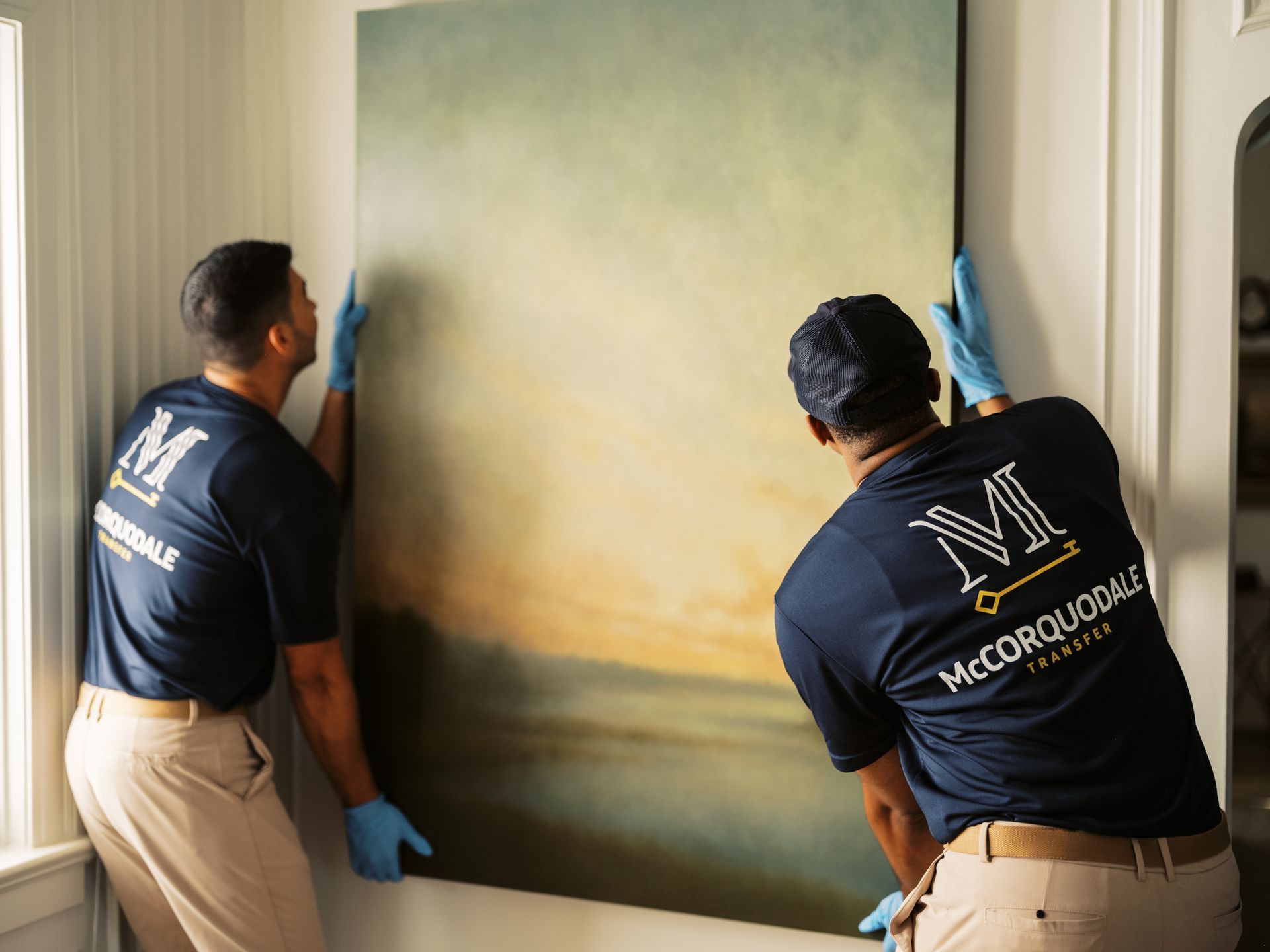 Two people in blue shirts and gloves hanging a large painting on a white wall.
