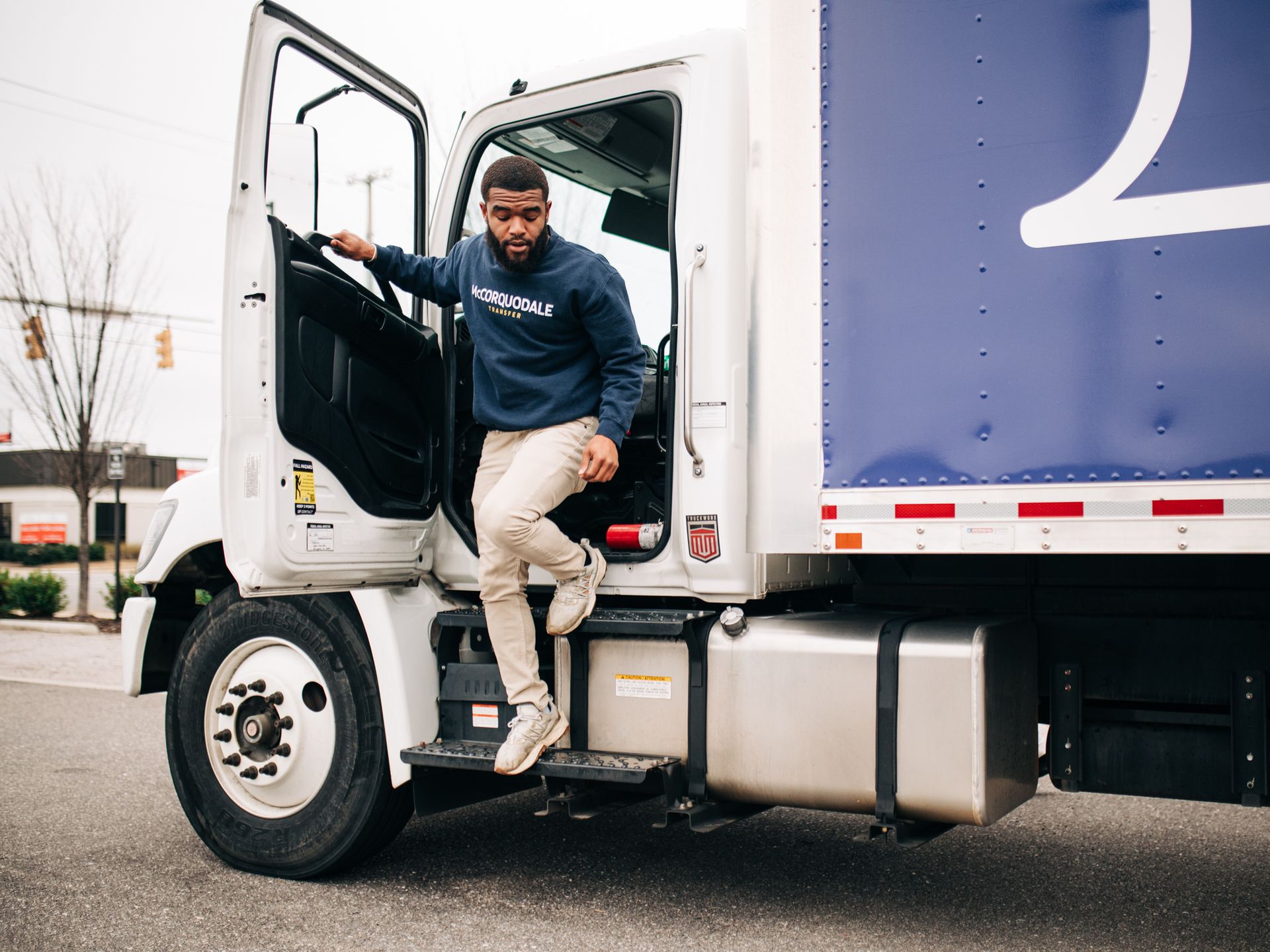 Person exiting a white truck with a blue cargo section. They wear a blue sweatshirt and tan pants.