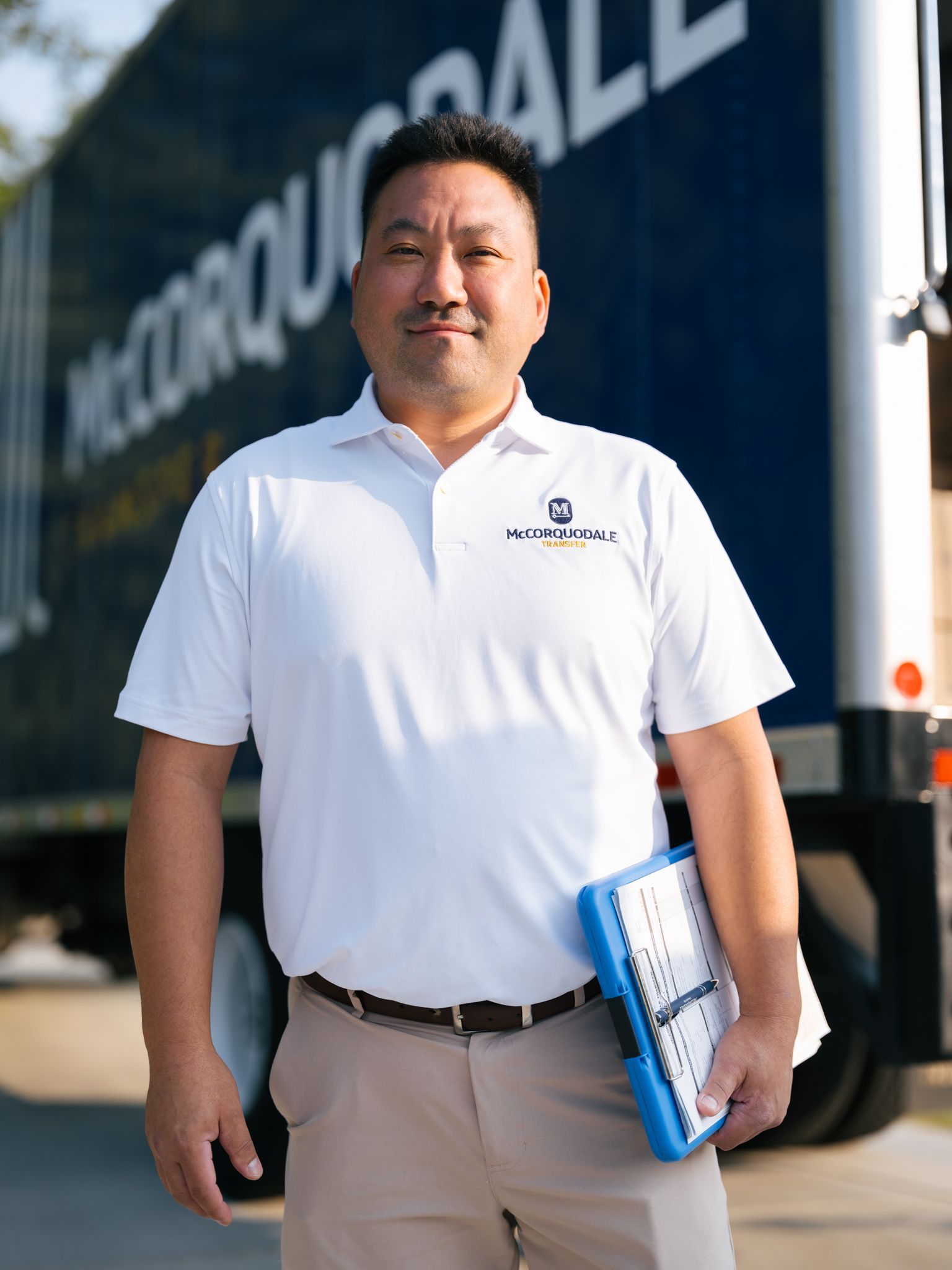 Man in white polo shirt holds clipboard, standing in front of a blue and white moving truck.
