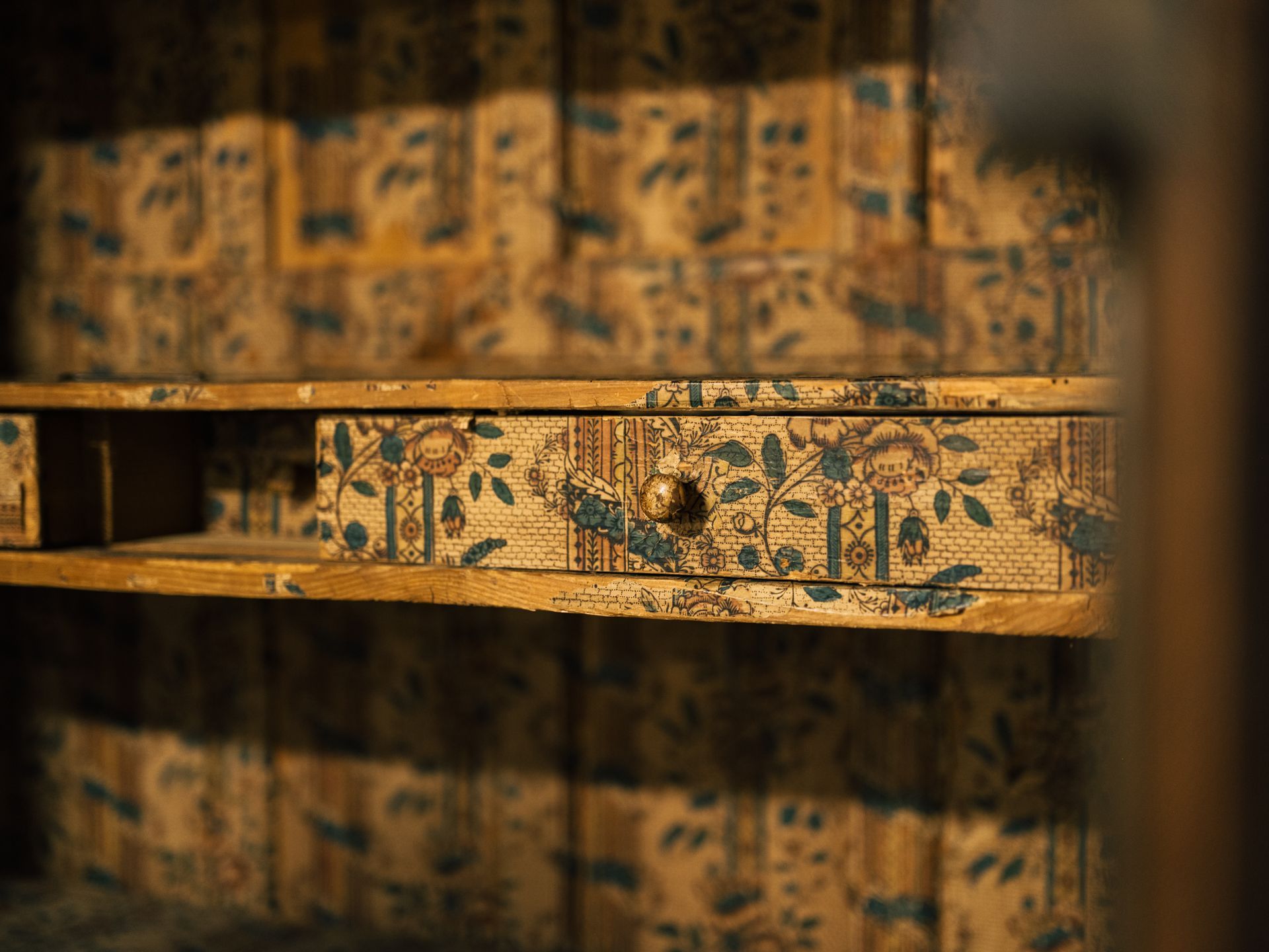 Interior of a wooden cabinet with blue and gold floral wallpaper lining the walls and a drawer.