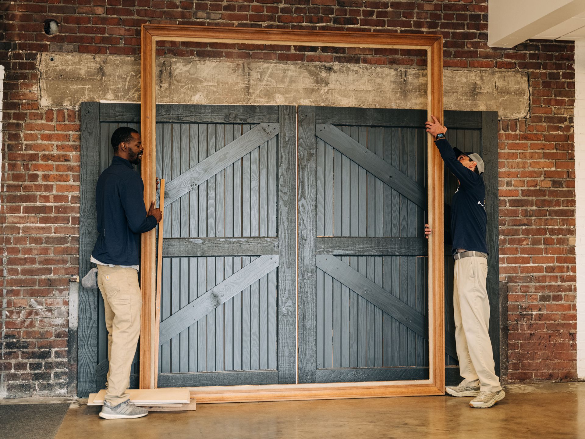 Two people hold a large wooden frame against a gray door and brick wall.