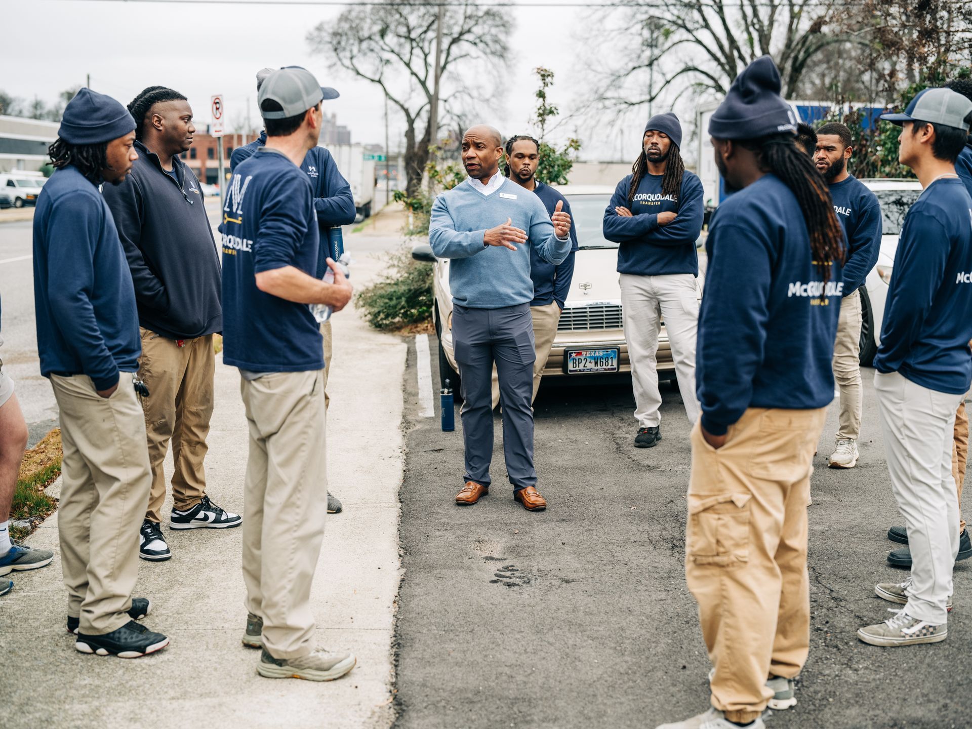 A group of people in blue uniforms listens to a man in a light blue sweater and khakis speaking outdoors.