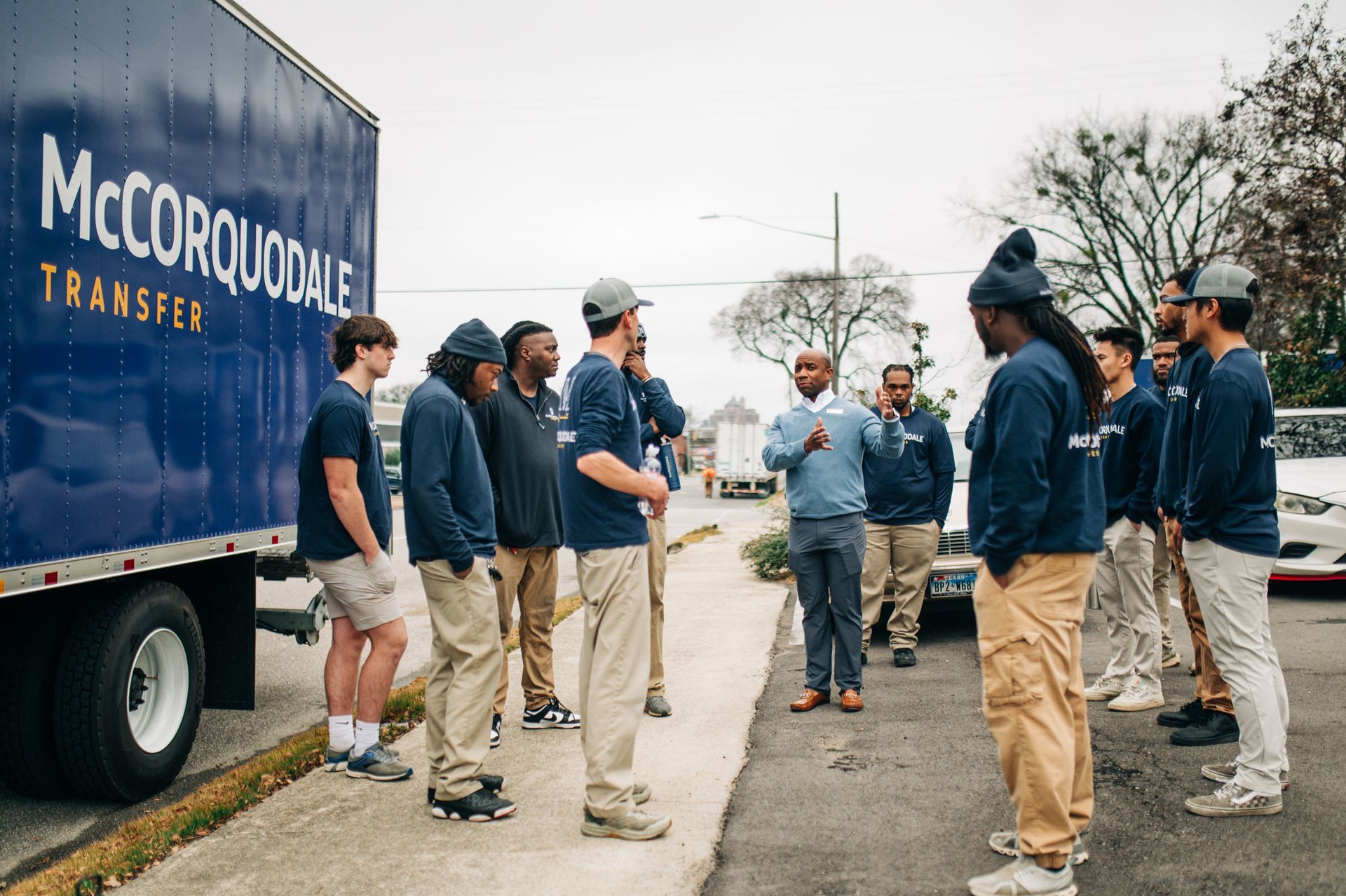 Movers in uniform stand around a truck, listening to a man speaking, outdoors.