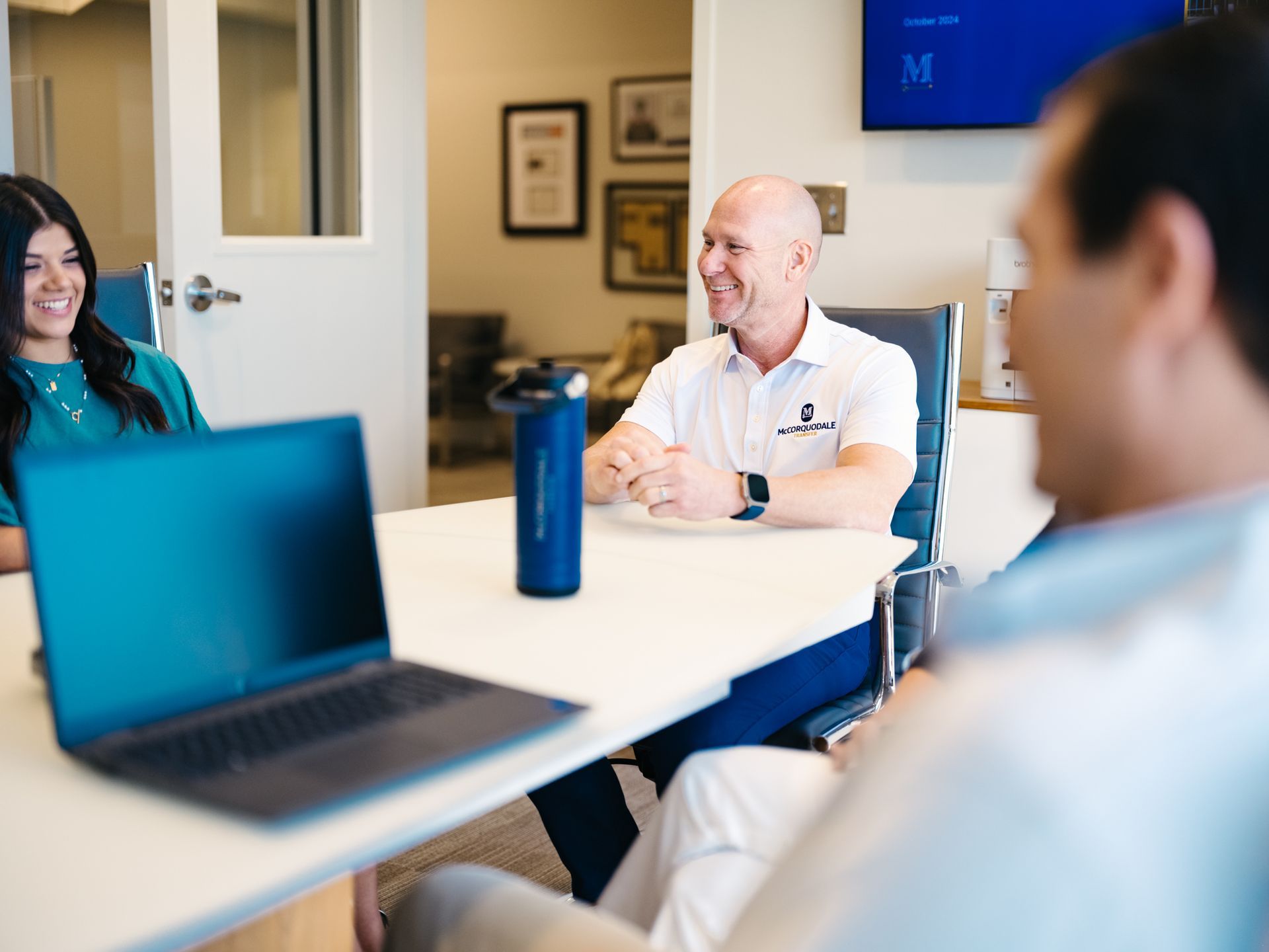 People in meeting: woman, man smiling, laptop, water bottle on white table.