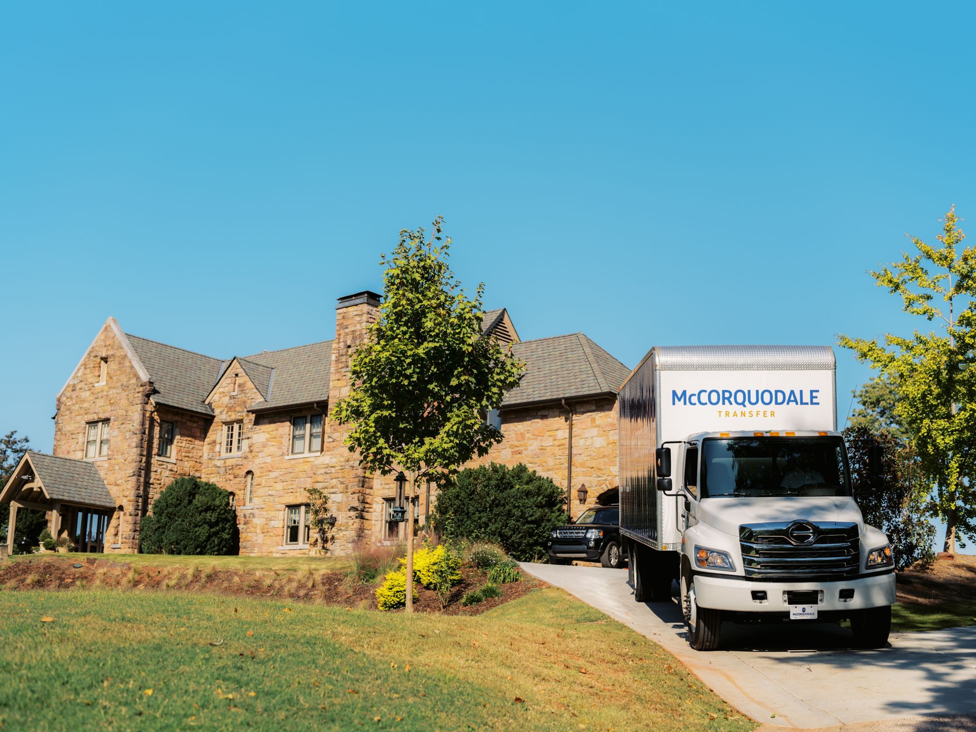 Moving truck in driveway of large stone house under blue sky.
