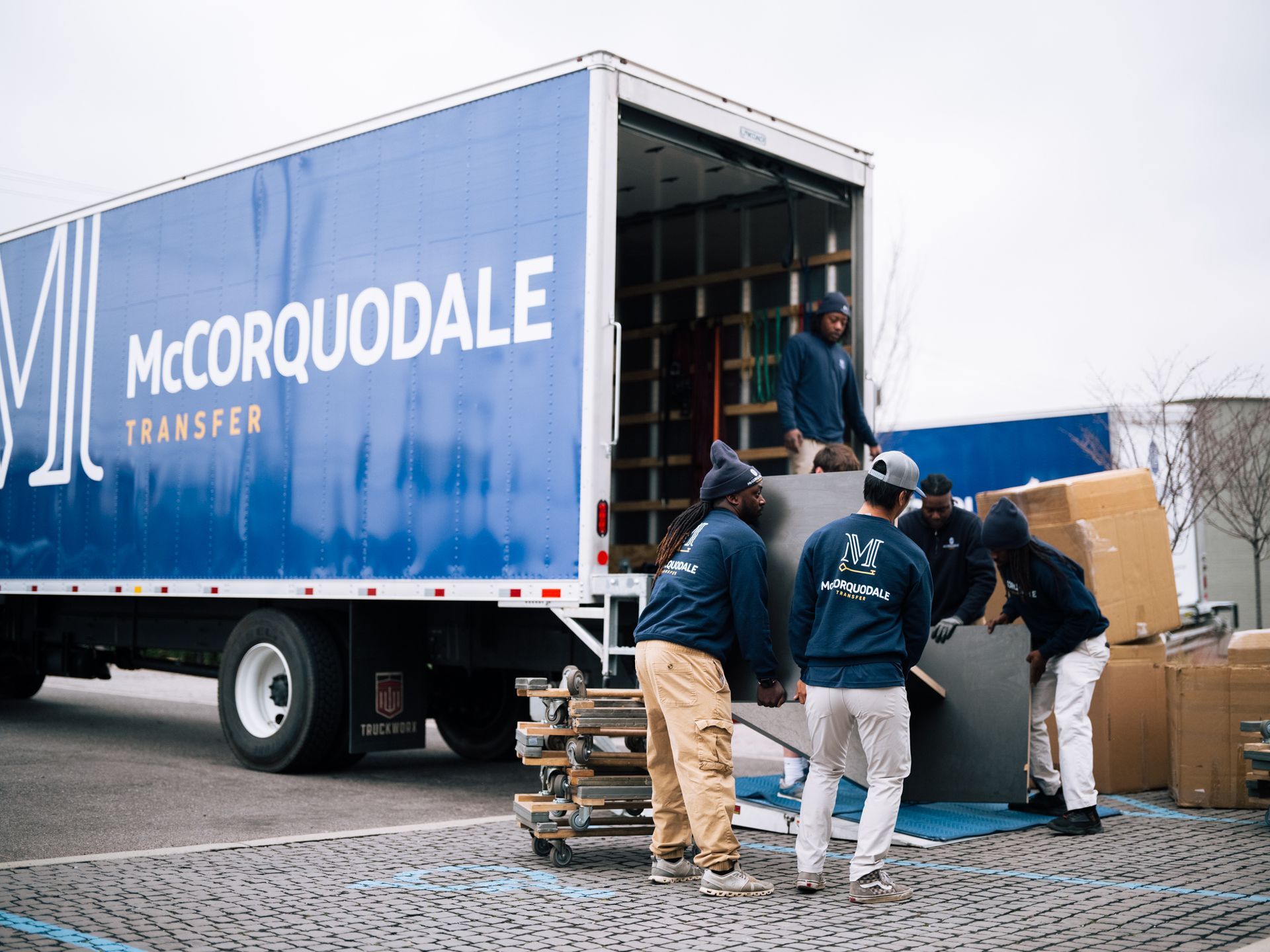 Movers loading a large truck with blue and white branding; carrying furniture. Outdoors, overcast sky.