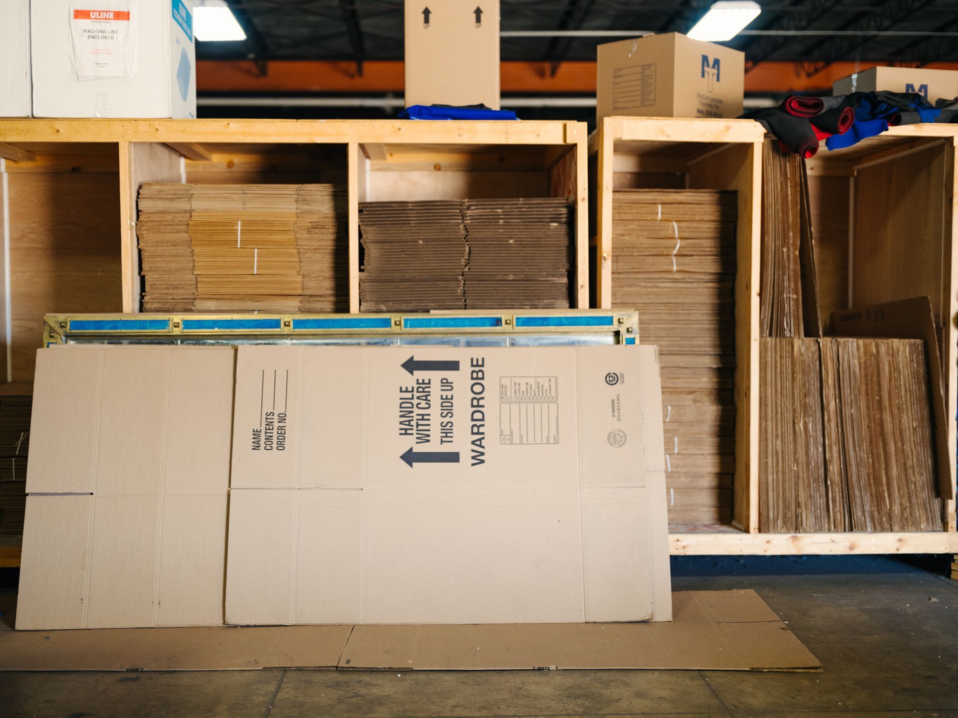 Stacks of flattened cardboard boxes in a warehouse, some in wooden storage units.