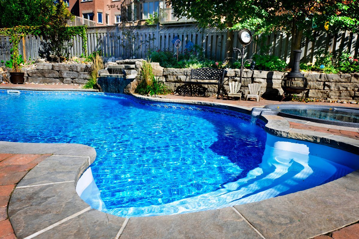 Blue swimming pool with steps, surrounded by stone and trees in a backyard.