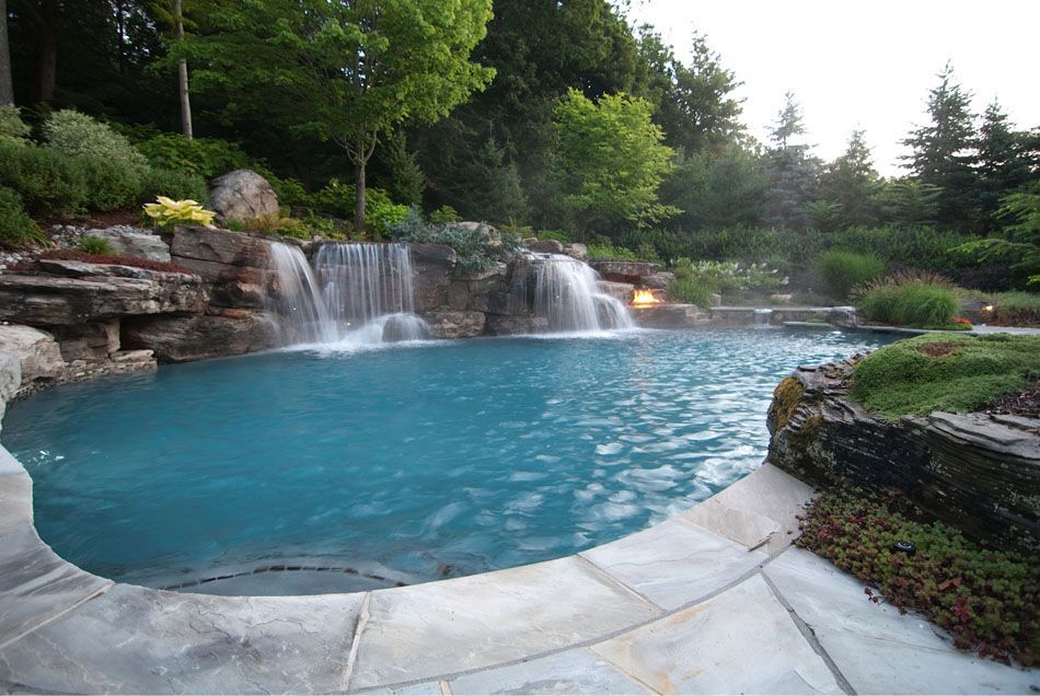 A backyard pool with a waterfall feature surrounded by lush greenery and stone.