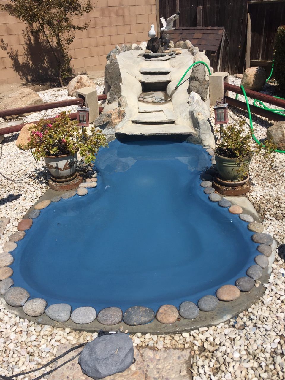 Pond with blue liner, waterfall, and rock border, in a yard with gravel and potted plants.