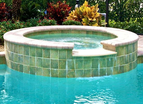 A circular hot tub with green tiles integrated into a turquoise pool, surrounded by landscaping.