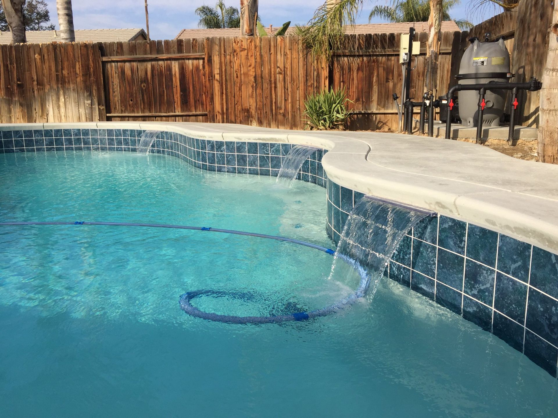 Pool with blue tile and cascading water, concrete border, wooden fence in the background.
