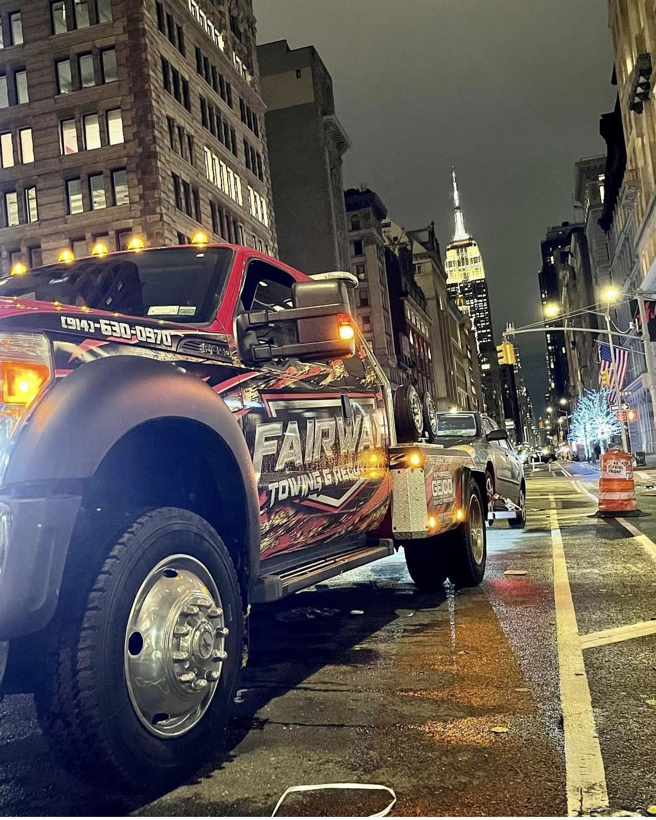 Tow truck parked on a wet city street at night, with the Empire State Building in the background. The truck has colorful graphics and bright lights.