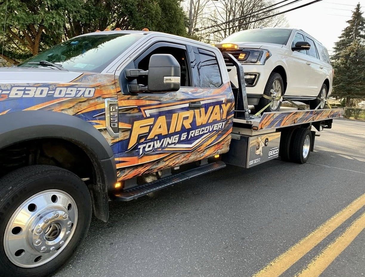 A white SUV being towed by a Fairway Towing & Recovery truck on a road with yellow lines.
