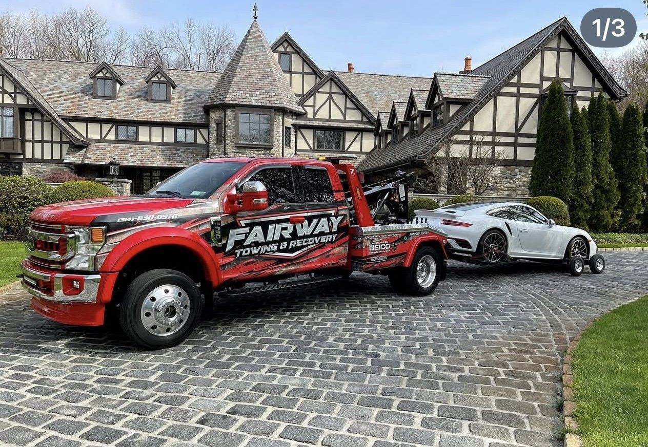 A red Fairway Towing truck towing a silver sports car out of a cobblestone driveway in front of a Tudor-style mansion.