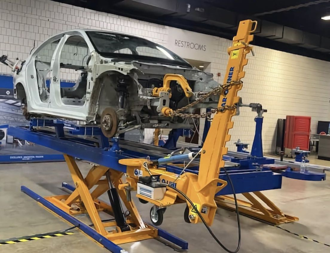 A car frame is undergoing repairs on a blue hydraulic lift, with a yellow pulling machine attached. The setting appears to be a car repair shop.