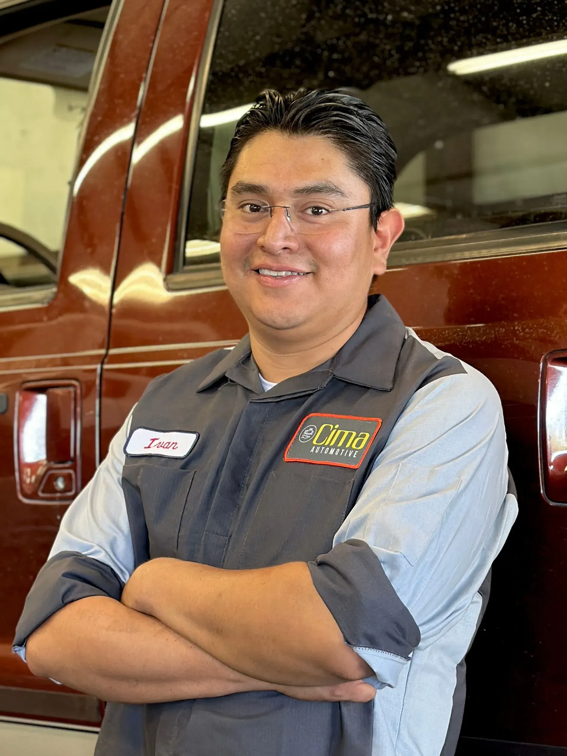 Mechanic with arms crossed, wearing a uniform. Leaning against a vehicle, smiling.