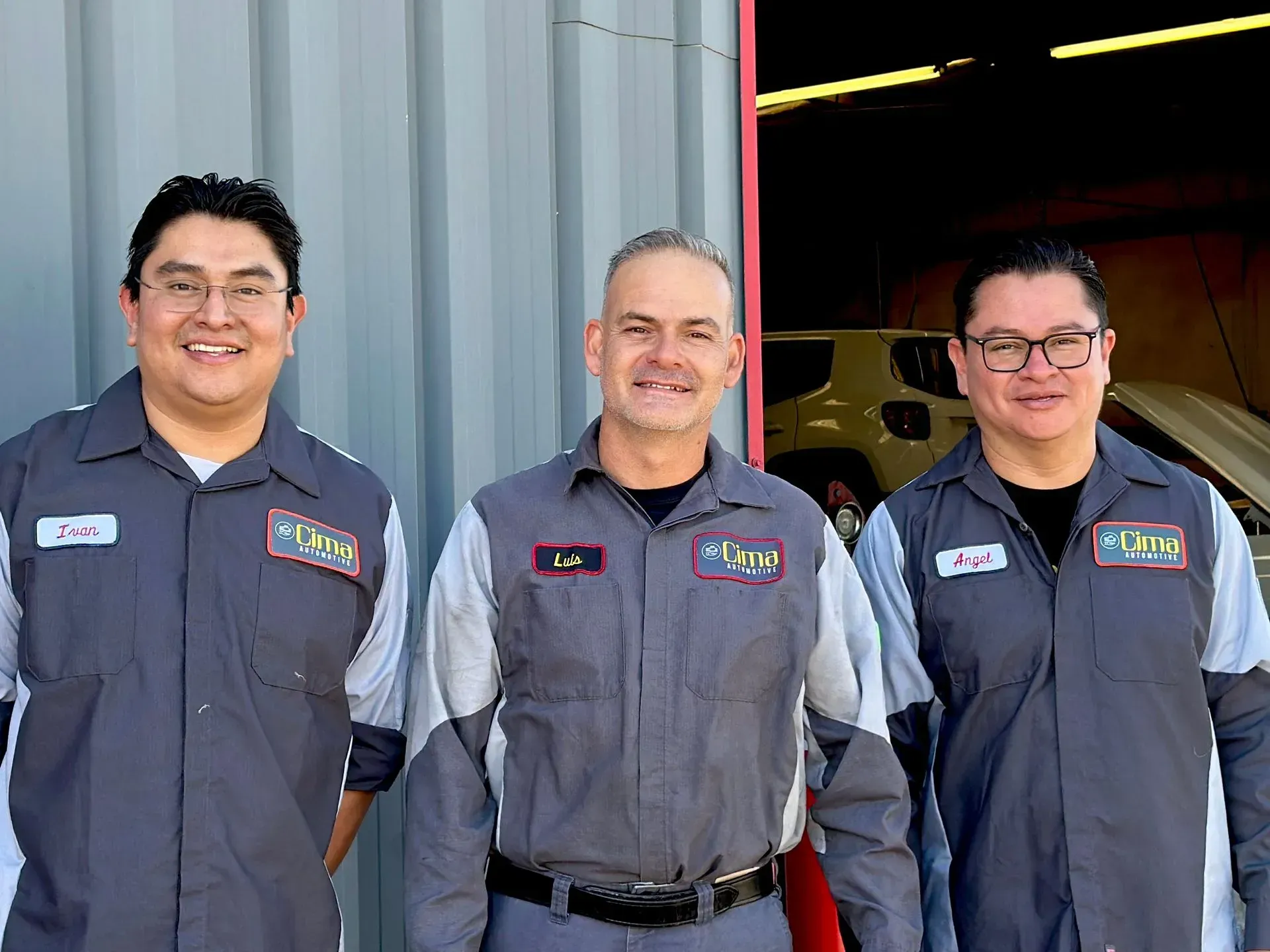 Three auto mechanics in grey work uniforms stand outside a garage. Two men smile, the middle man has a neutral expression.